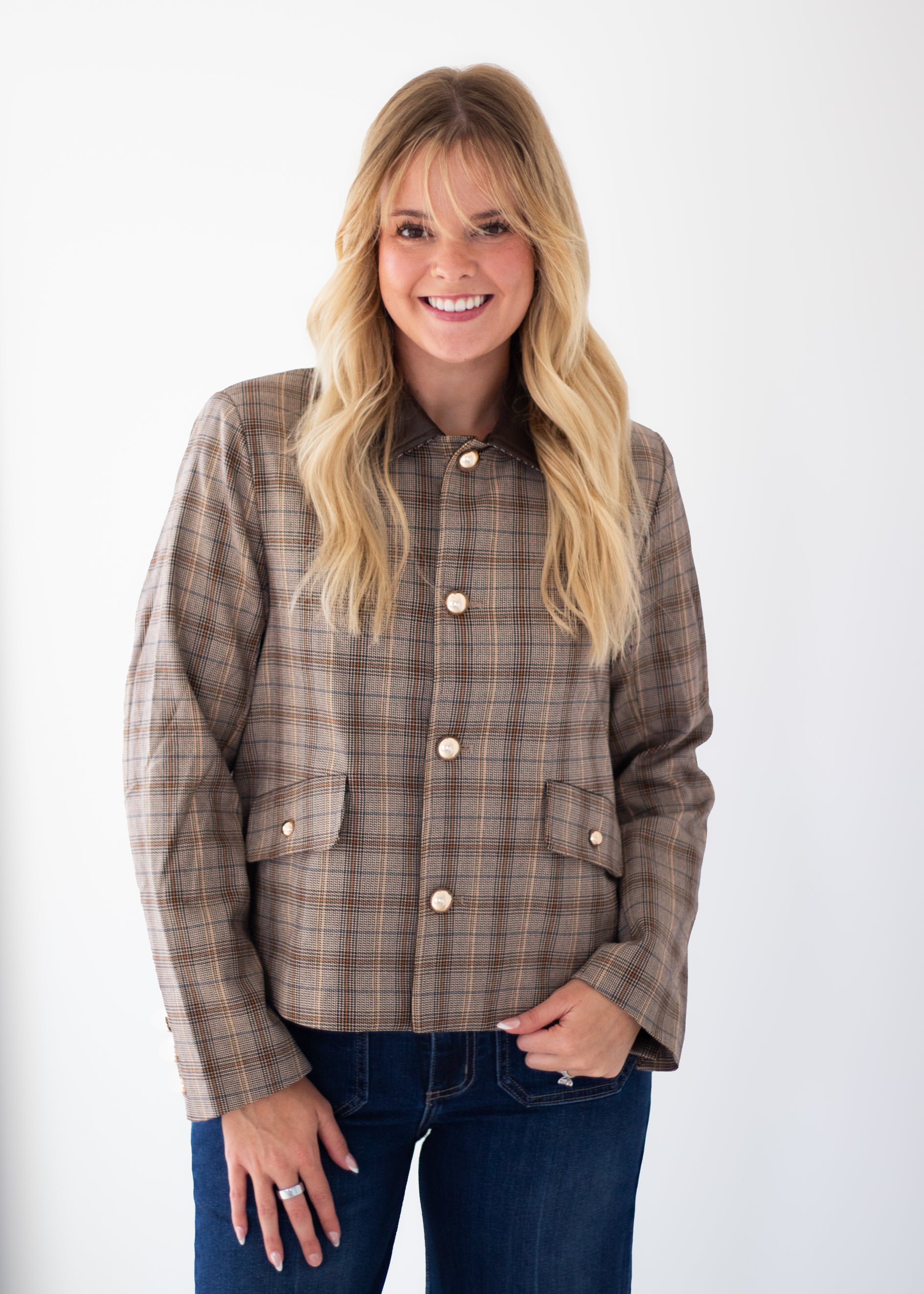 A smiling blonde woman wearing a brown and beige plaid button-front jacket with flap pockets, paired with dark blue jeans, standing against a plain white studio background.
