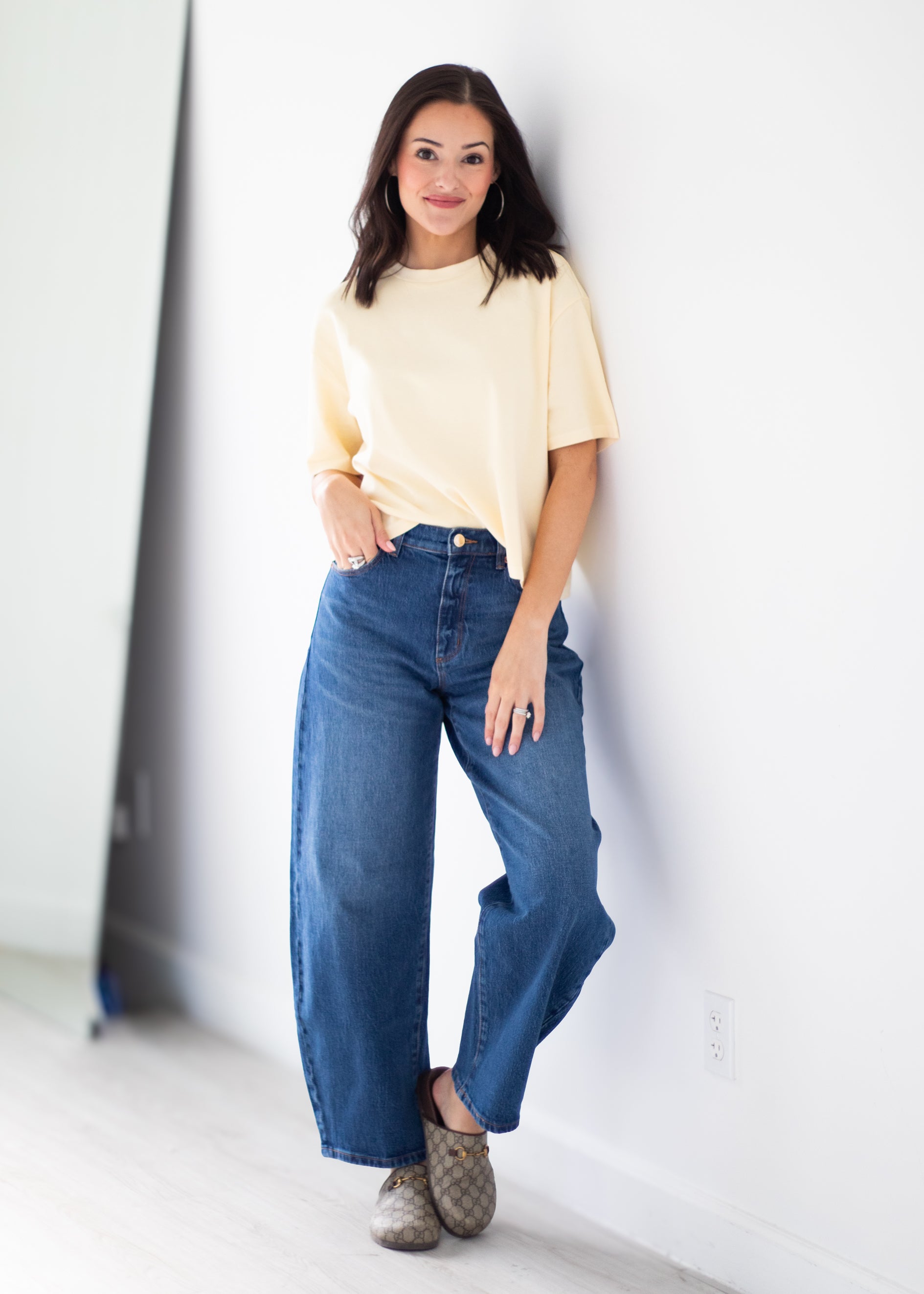 A woman stands against a white wall wearing a pale yellow short-sleeve top, wide-leg blue jeans, and brown patterned slip-on shoes.