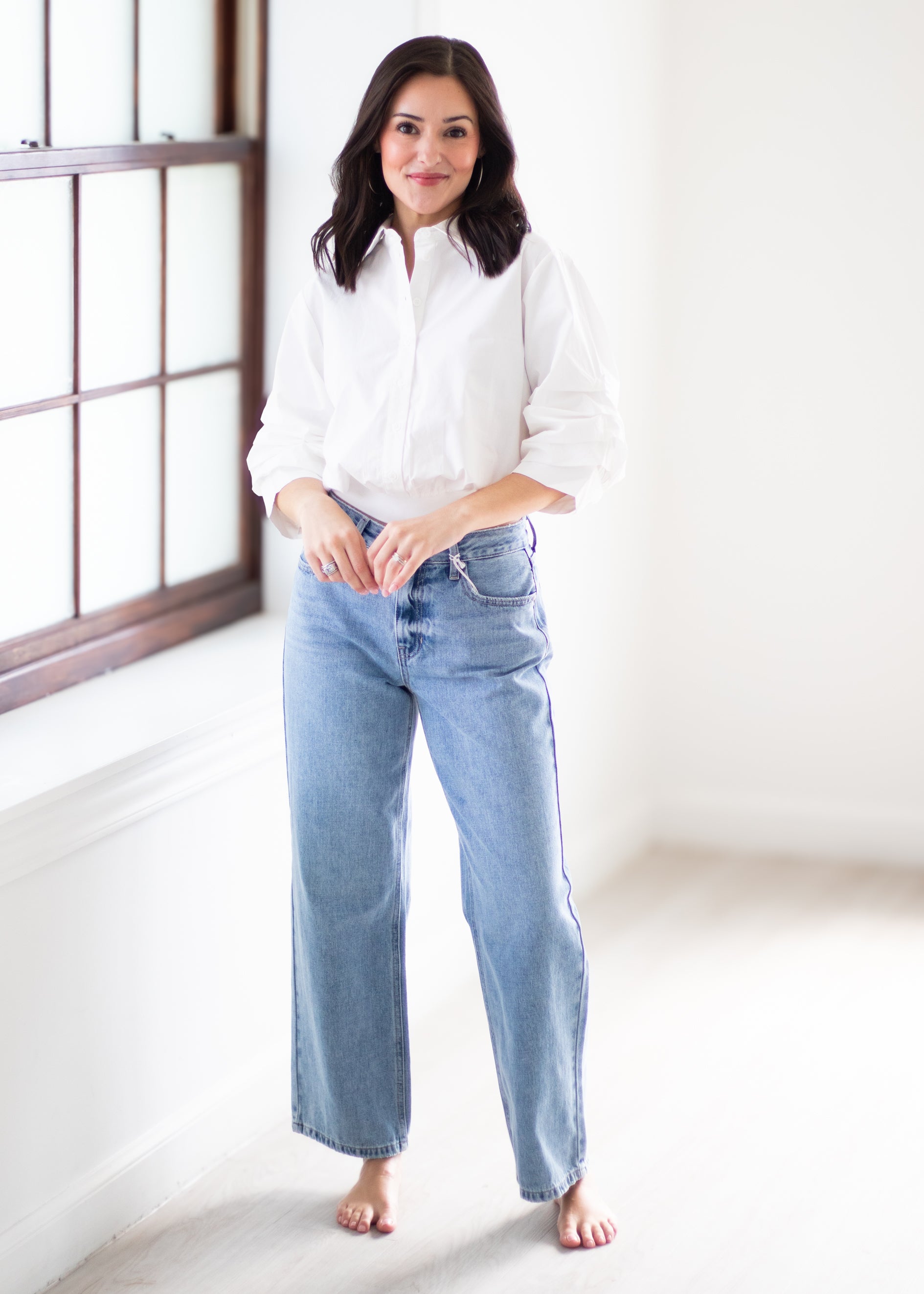 A woman stands barefoot in a bright room wearing a white button-up shirt and light-wash blue wide-leg jeans.