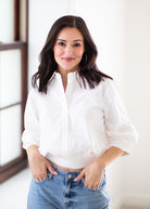 A woman with dark hair wearing a white button-up shirt and light blue jeans, standing in a brightly lit indoor setting.