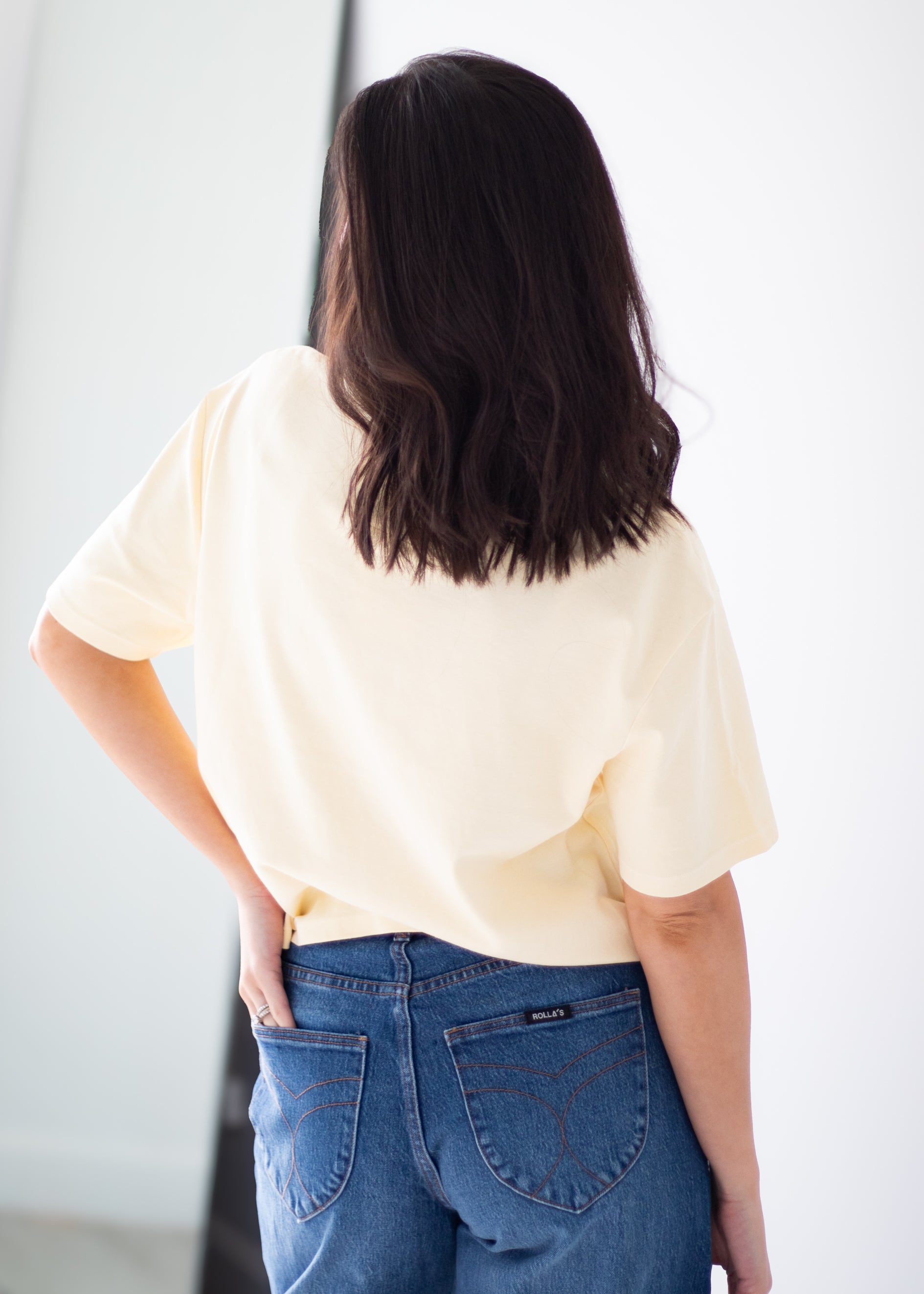 A woman viewed from behind wearing a pale cream short-sleeve t-shirt and blue jeans, standing against a bright white background.