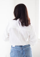 A woman viewed from behind wearing a white long-sleeve blouse with a gathered back neckline and light blue jeans, standing against a bright white indoor background.