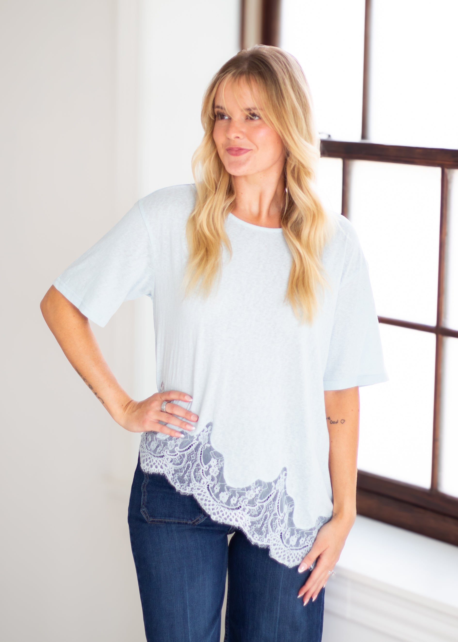 A woman modeling a light blue short-sleeve top with a white lace hem, paired with dark blue jeans, standing in a brightly lit room with a window.