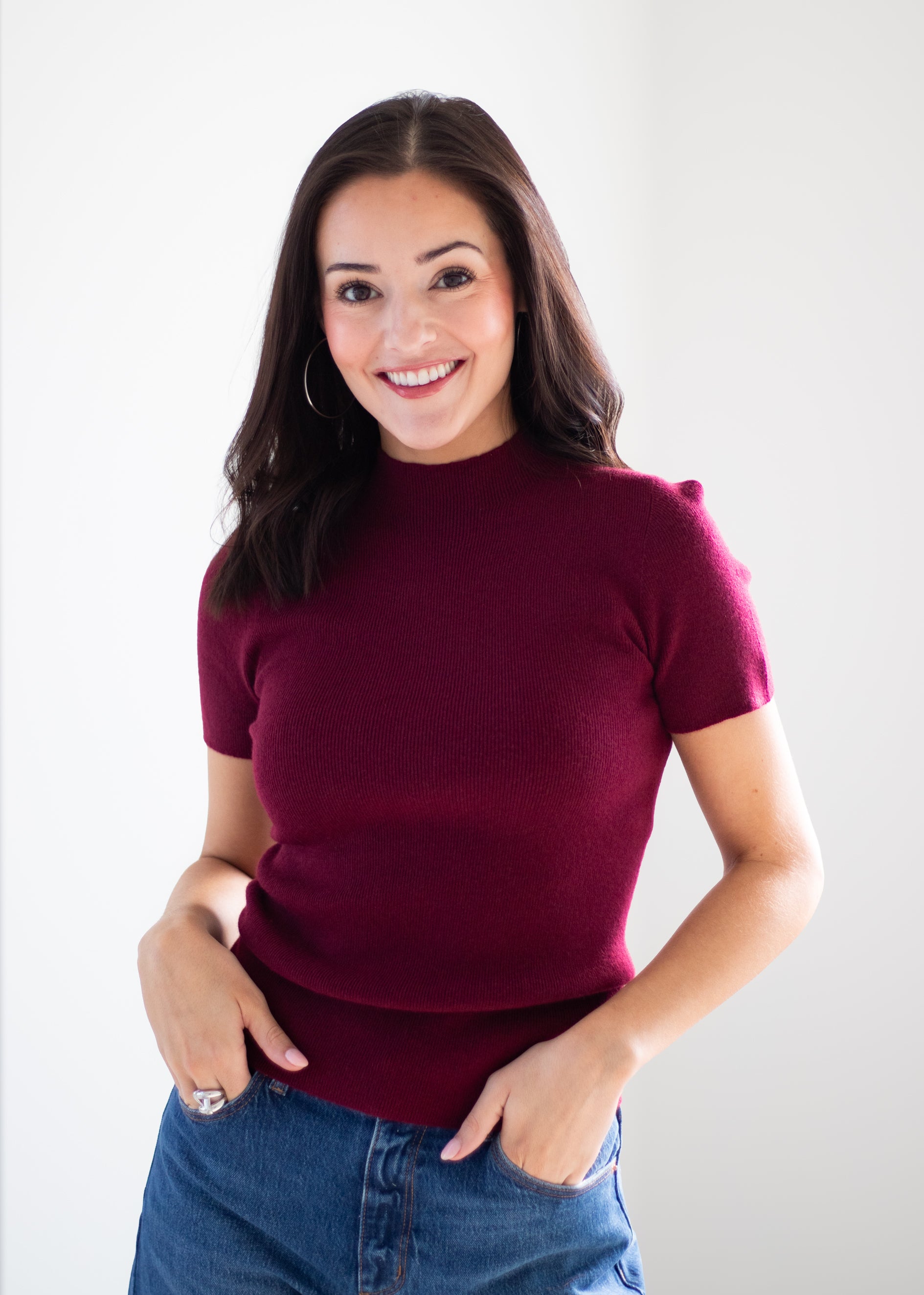 A woman with long dark brown hair smiling, wearing a short-sleeve mock neck knit top in deep burgundy and blue jeans, standing against a light gray studio background.
