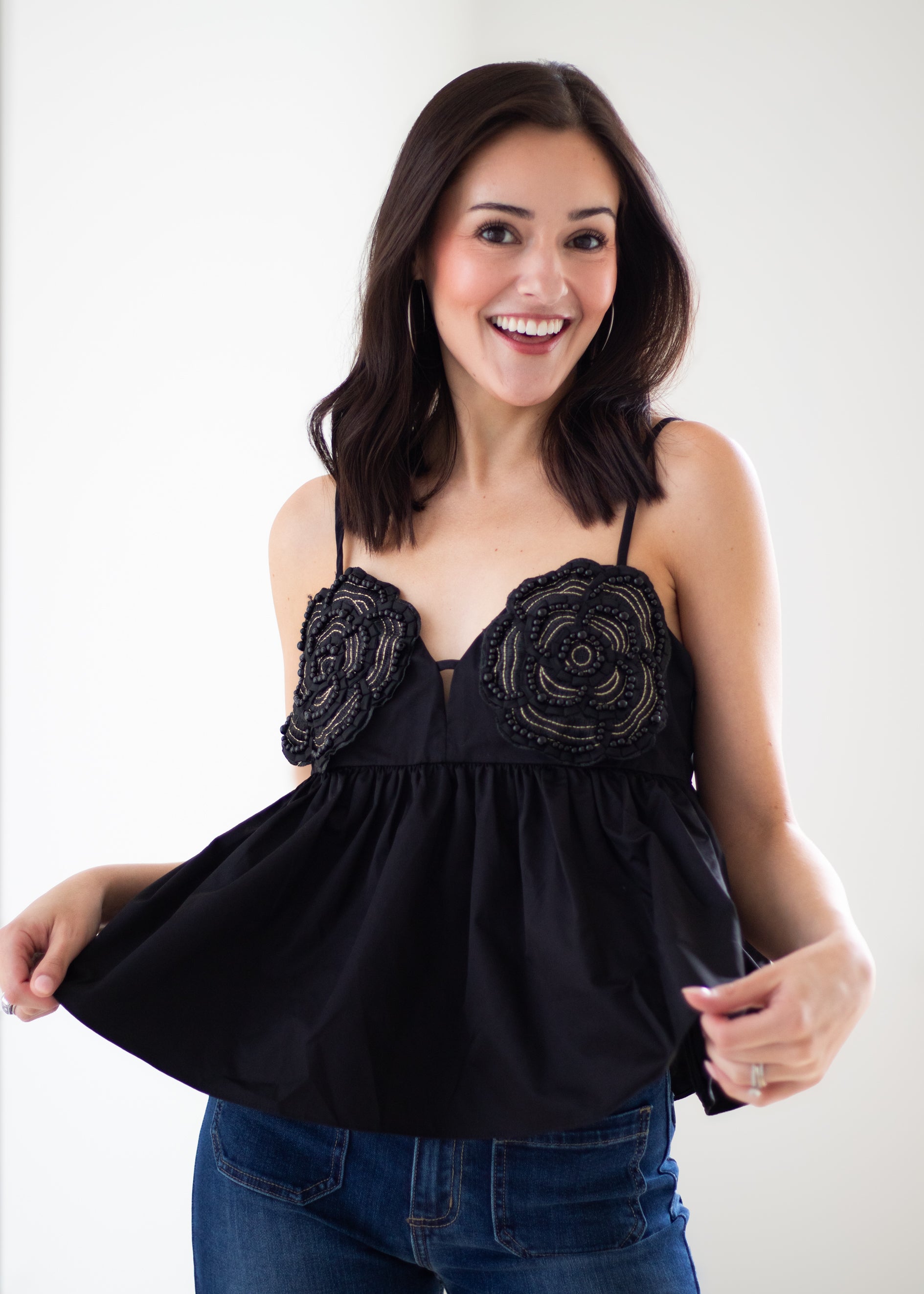 A smiling woman wearing a black spaghetti-strap top with 3D floral applique on the bust and a flared peplum skirt, paired with blue jeans, against a plain white studio background.