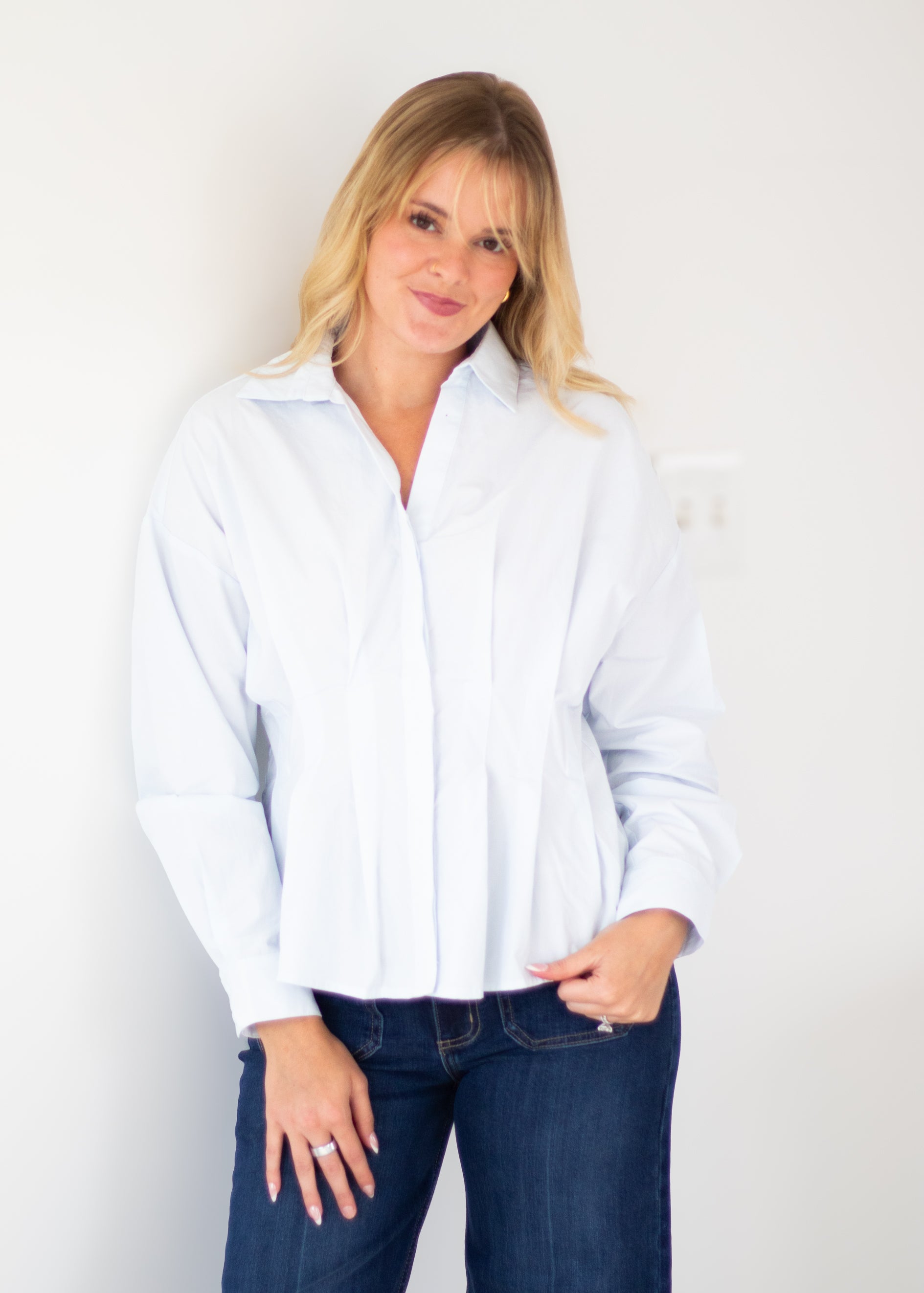 A woman with blonde hair wearing a light blue long-sleeve button-down shirt and dark blue jeans, standing against a plain white wall.