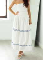 Woman wearing a white strapless dress with blue embroidery in a minimal indoor setting.