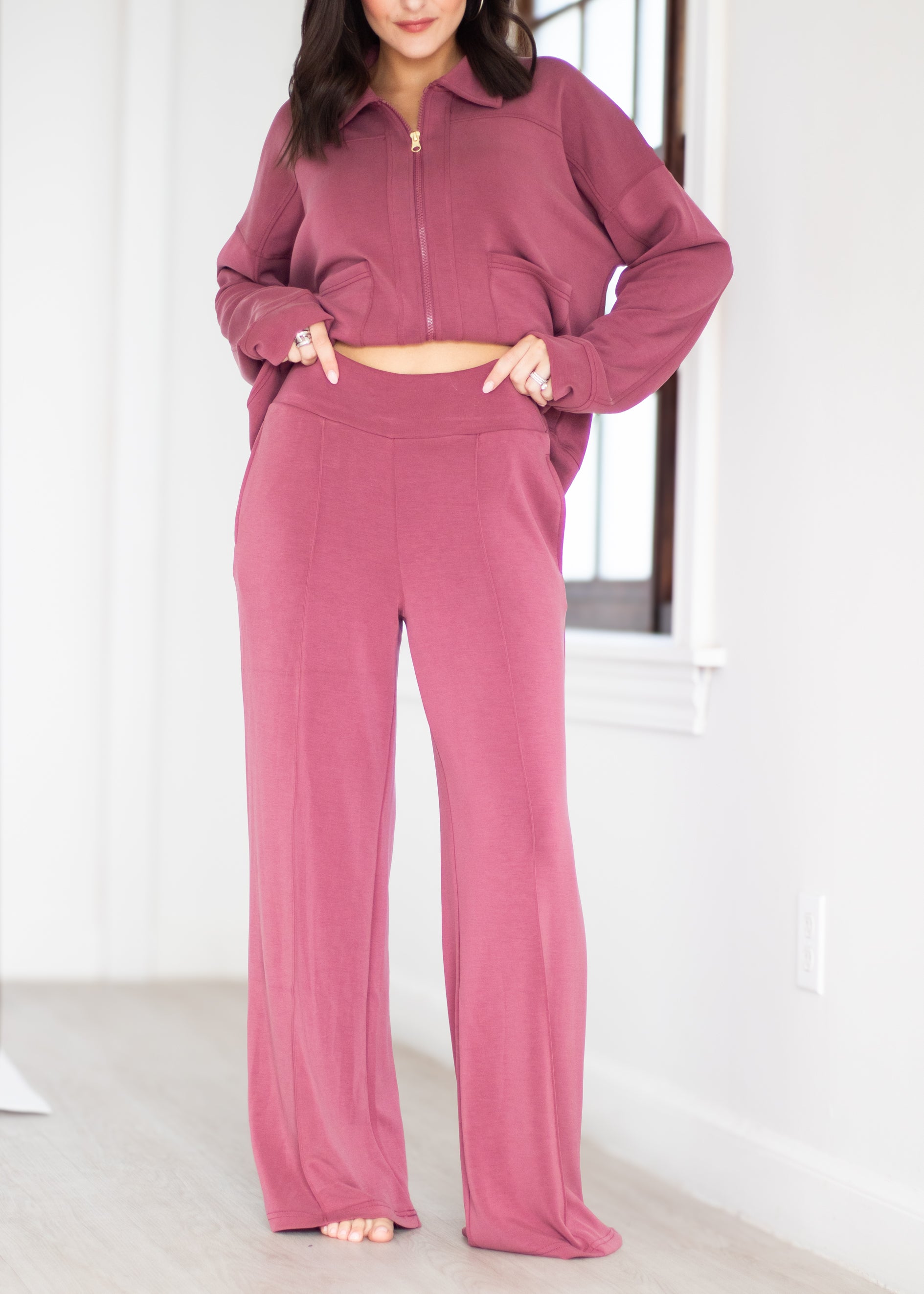 A woman modeling a matching dusty rose two-piece loungewear set consisting of a cropped zip-up jacket and high-waisted wide-leg pants, standing in a bright room.
