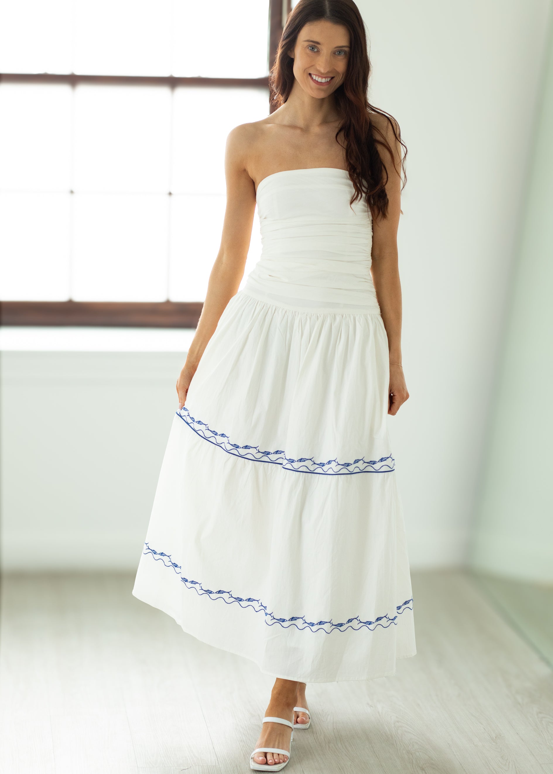 Woman wearing a white strapless dress with blue trim in a bright room.