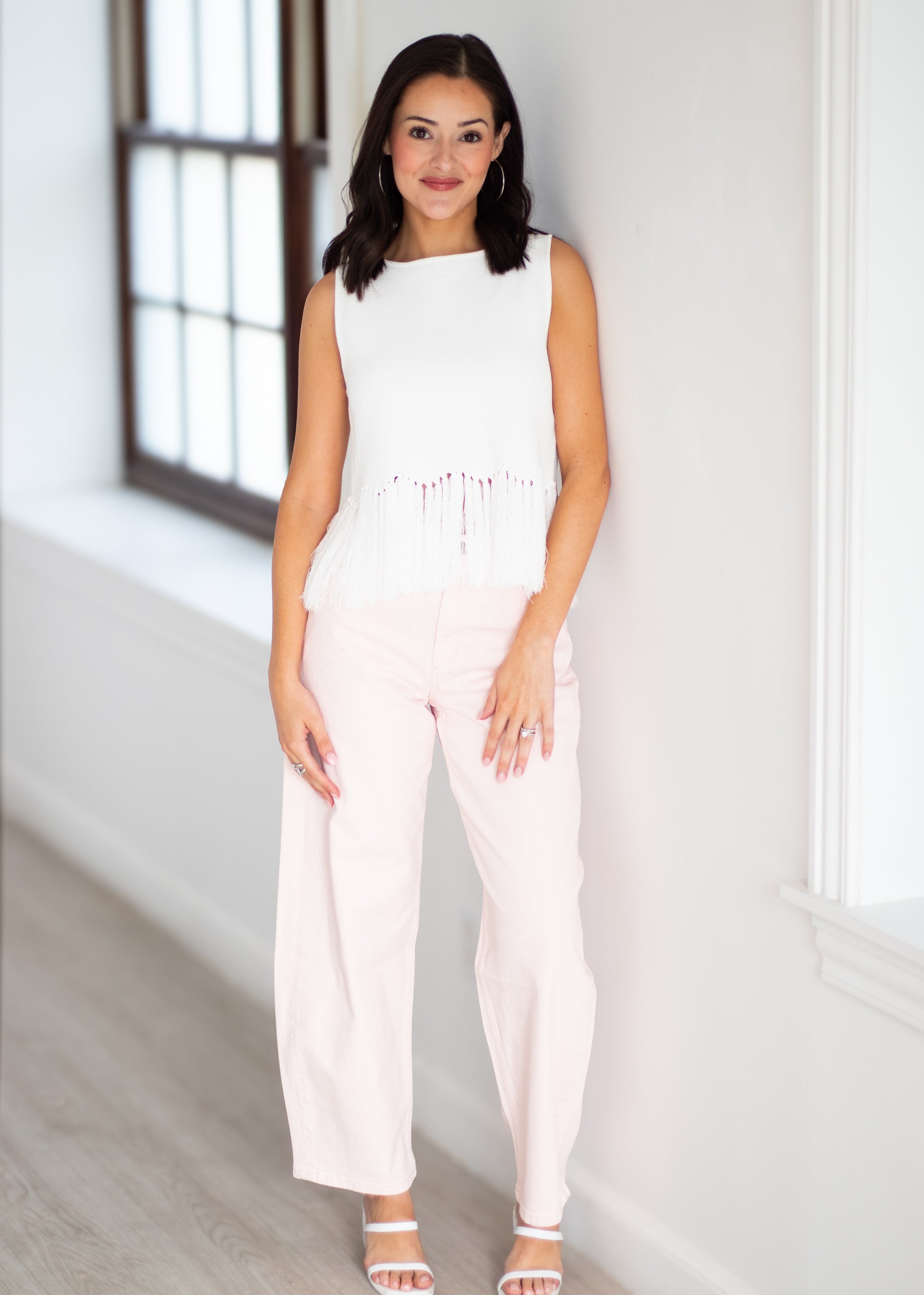 A woman modeling a sleeveless white top with fringe detailing and light pink wide-leg pants, standing in a bright indoor setting with white walls and a window.