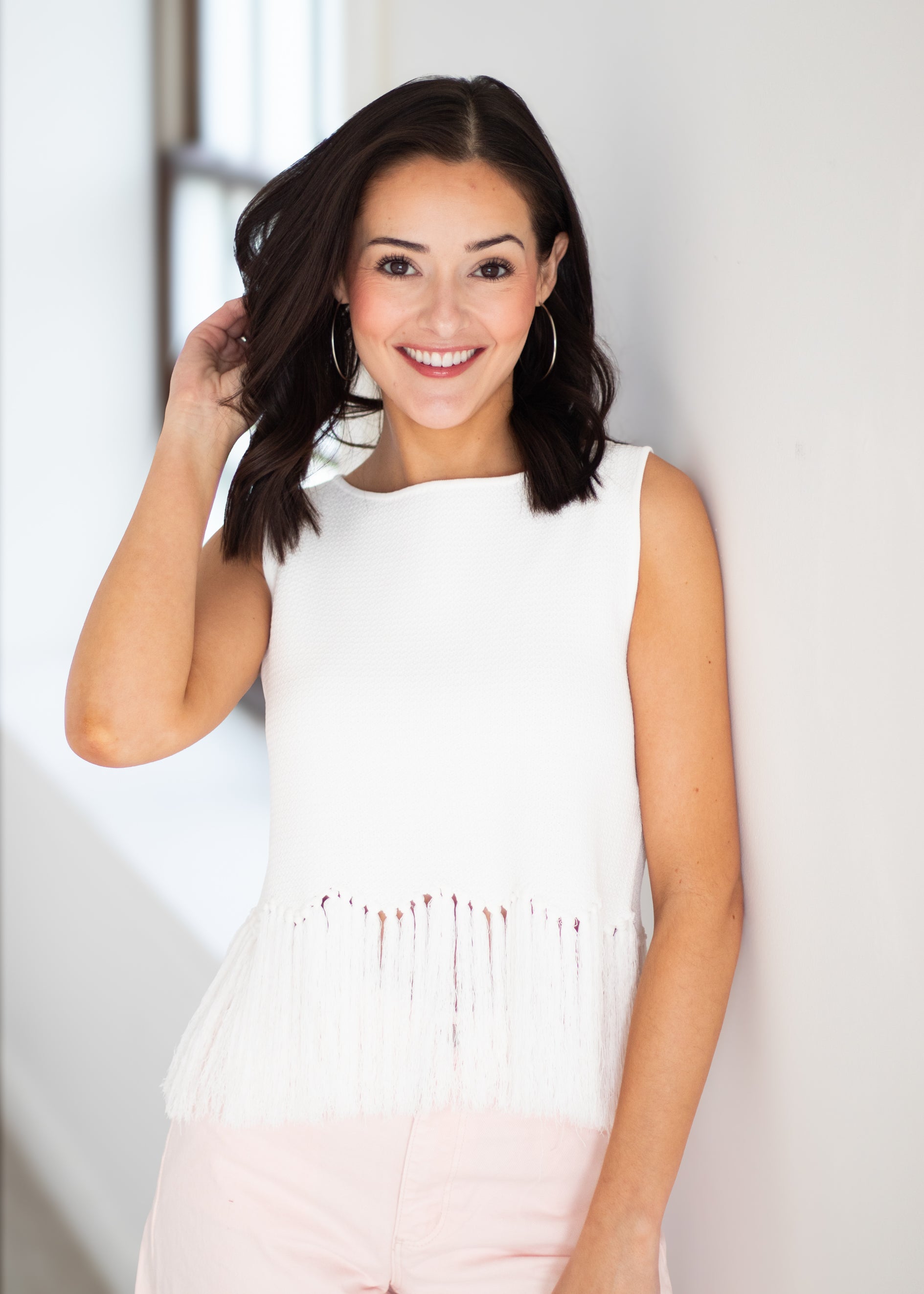 A woman with dark shoulder-length hair smiling and posing in a sleeveless white top with fringe detailing at the hem, paired with light pink pants, set against a bright white wall.