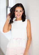 A woman with dark shoulder-length hair smiling and posing in a sleeveless white top with fringe detailing at the hem, paired with light pink pants, set against a bright white wall.