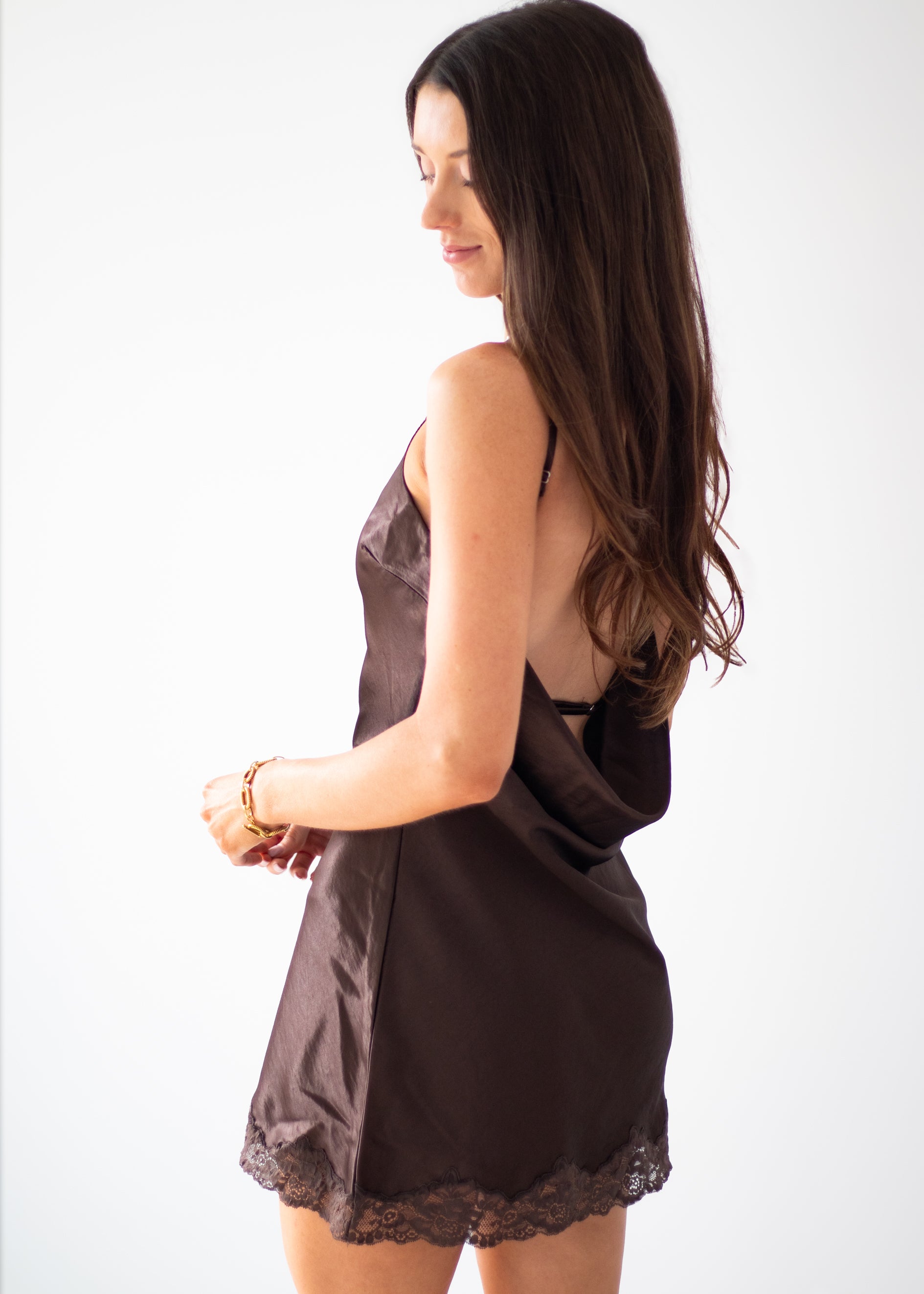 A woman with long dark brown hair is shown from the back wearing a dark brown satin slip dress with lace trim at the hem, set against a plain white studio background.