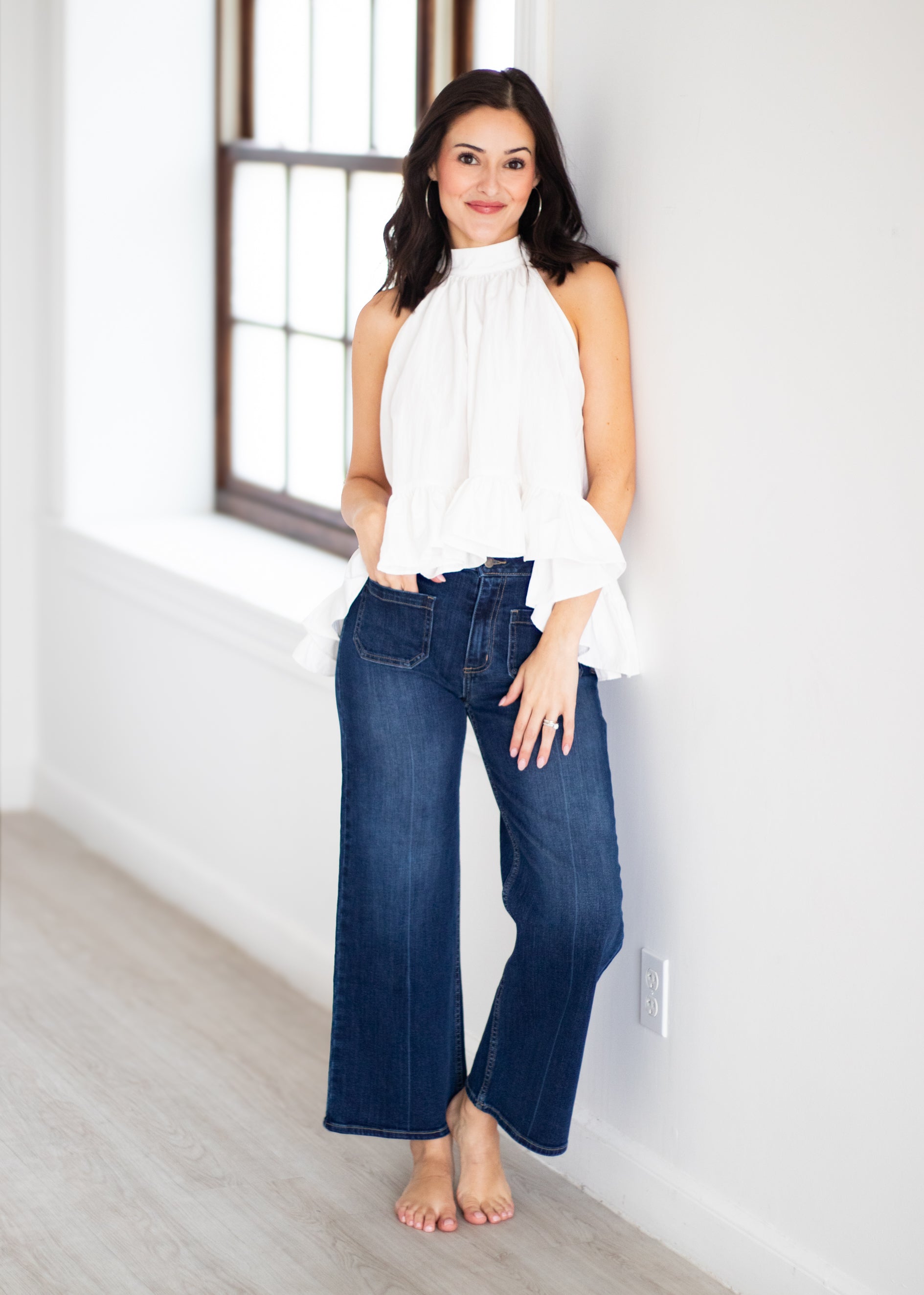 A woman stands barefoot in a bright room, wearing a white sleeveless ruffled blouse and dark blue wide-leg jeans.