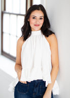A woman with dark hair wearing a white sleeveless high-neck blouse with ruffles and blue jeans, standing in a brightly lit room next to a window.