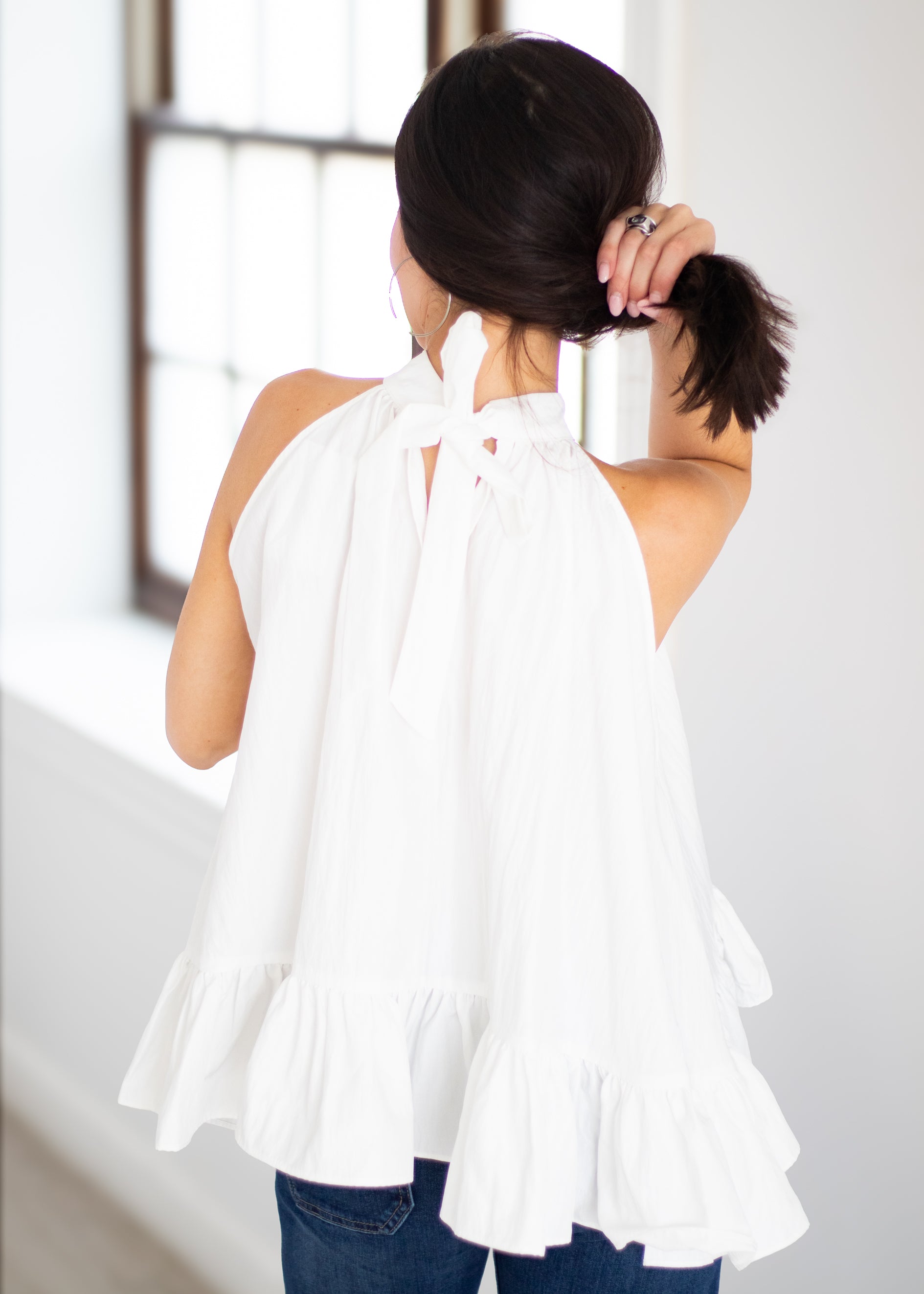 A woman shown from behind wearing a white sleeveless halter top with a tie-back and ruffled hem, paired with blue jeans in a bright indoor setting.