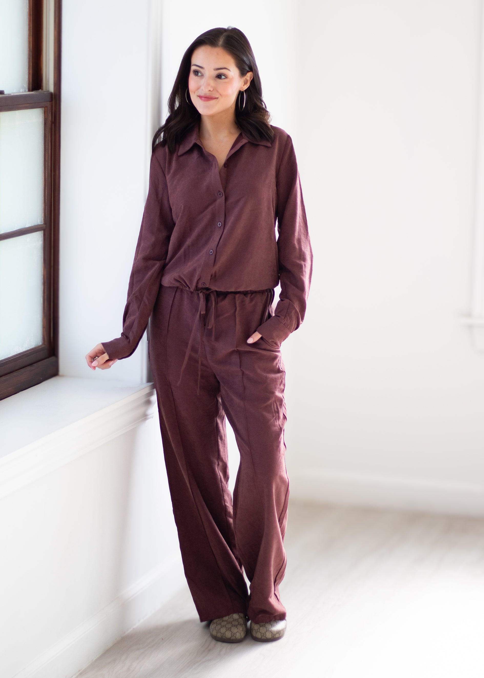A woman modeling a matching two-piece loungewear set in a dusty plum-brown color, consisting of a long-sleeve button-up shirt and wide-leg pants, standing in a bright, minimalist room.