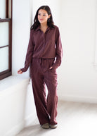 A woman modeling a matching two-piece loungewear set in a dusty plum-brown color, consisting of a long-sleeve button-up shirt and wide-leg pants, standing in a bright, minimalist room.