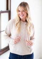 A smiling blonde woman wearing a beige textured knit zip-up hoodie sweater with drawstrings and front pockets, paired with blue jeans, standing in a brightly lit room.