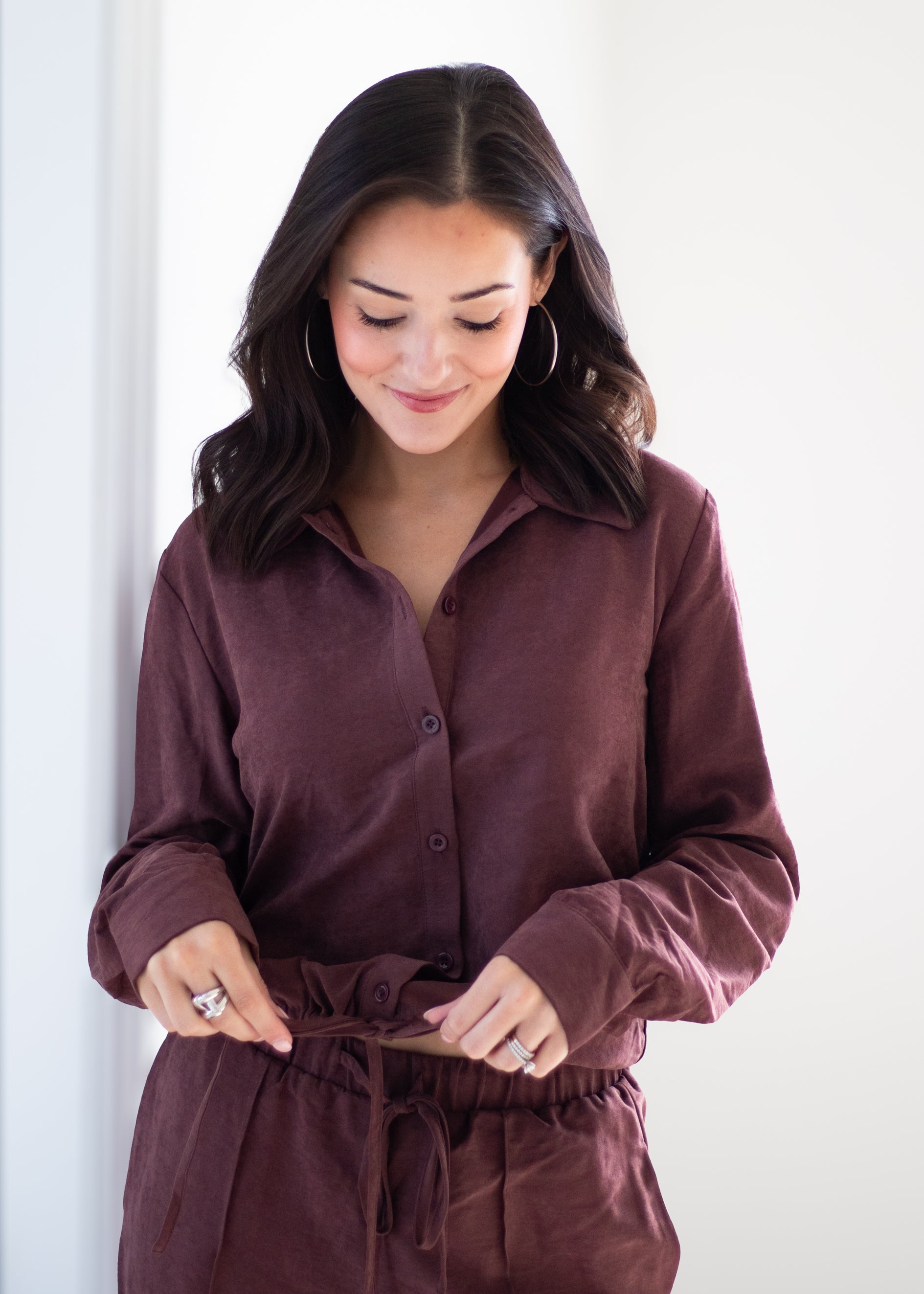 A woman wearing a matching dark plum button-up shirt and drawstring pants, looking down as she ties the waist. The photo is taken in a brightly lit, minimalist indoor setting.