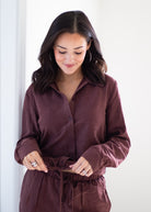A woman wearing a matching dark plum button-up shirt and drawstring pants, looking down as she ties the waist. The photo is taken in a brightly lit, minimalist indoor setting.