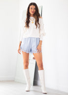 A woman modeling a white off-the-shoulder long-sleeve top, light blue lace-trimmed shorts, and white knee-high boots, standing in a bright white room with a large mirror behind her.