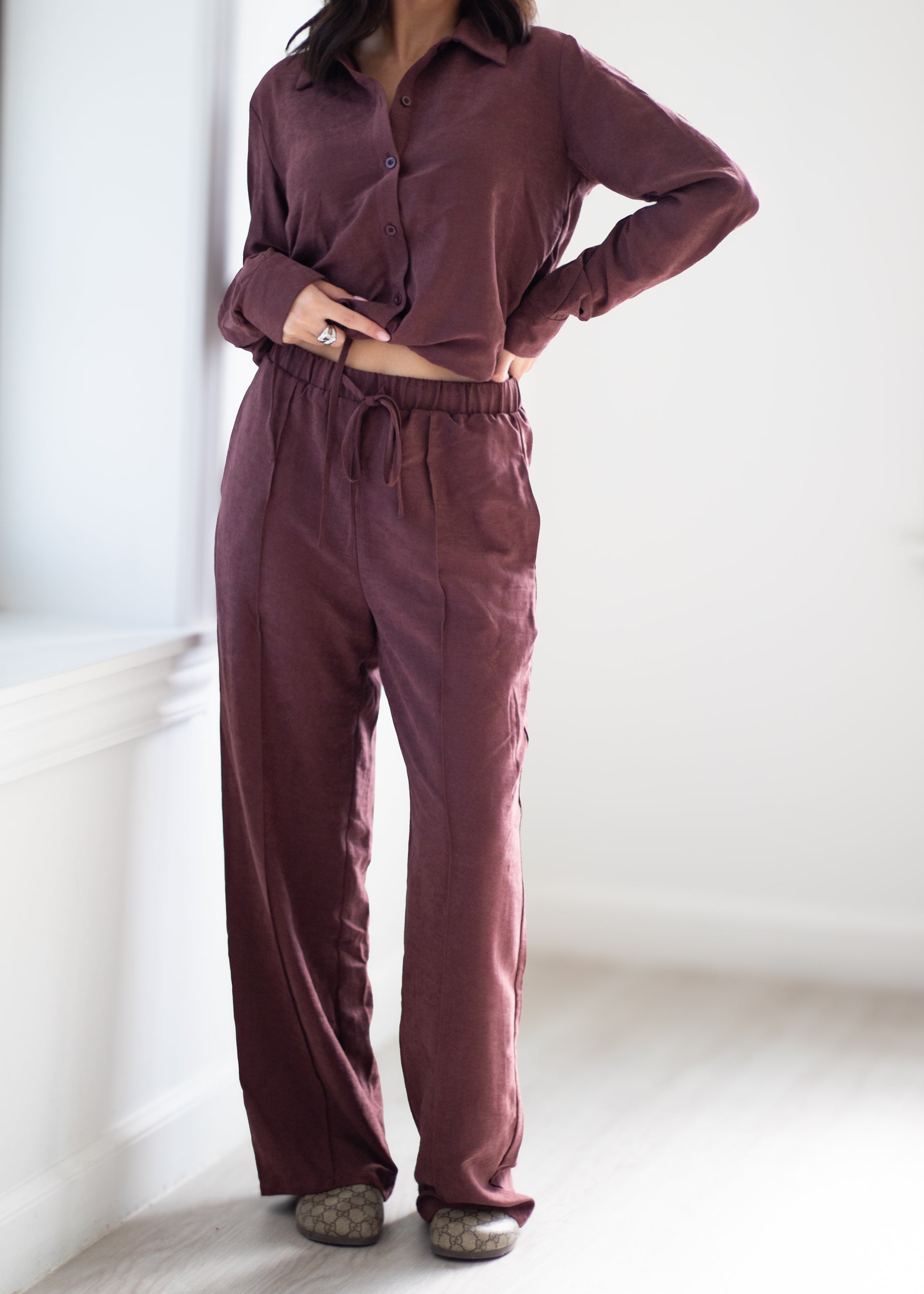 A person wearing a matching two-piece loungewear set in a dusty plum-brown color, featuring a long-sleeve button-up shirt and wide-leg drawstring pants, standing in a bright indoor room.