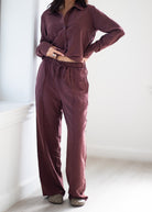 A person wearing a matching two-piece loungewear set in a dusty plum-brown color, featuring a long-sleeve button-up shirt and wide-leg drawstring pants, standing in a bright indoor room.