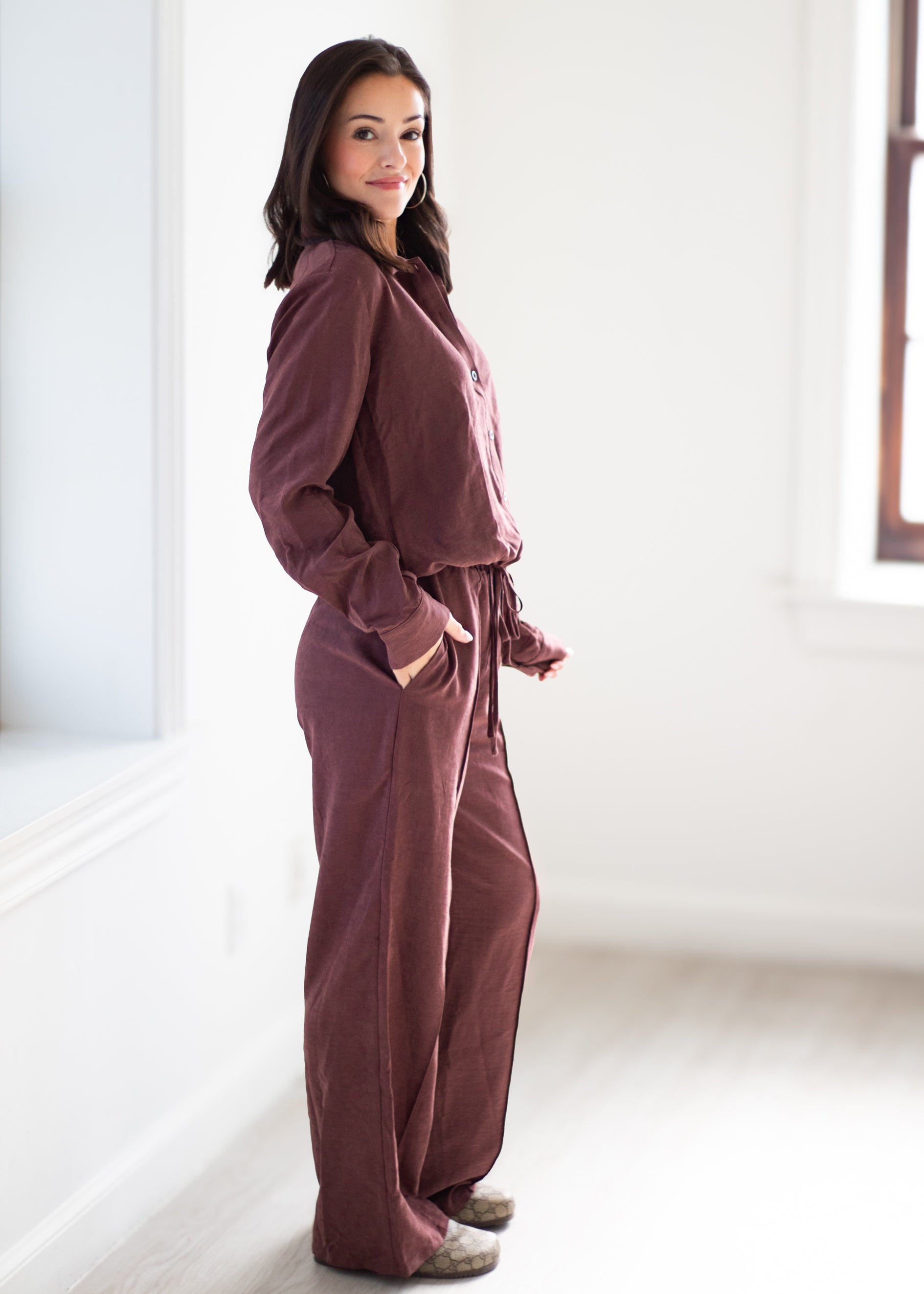 A woman wearing a matching mauve-brown lounge set consisting of a long-sleeve collared top and wide-leg pants, standing in a bright, minimalist room.
