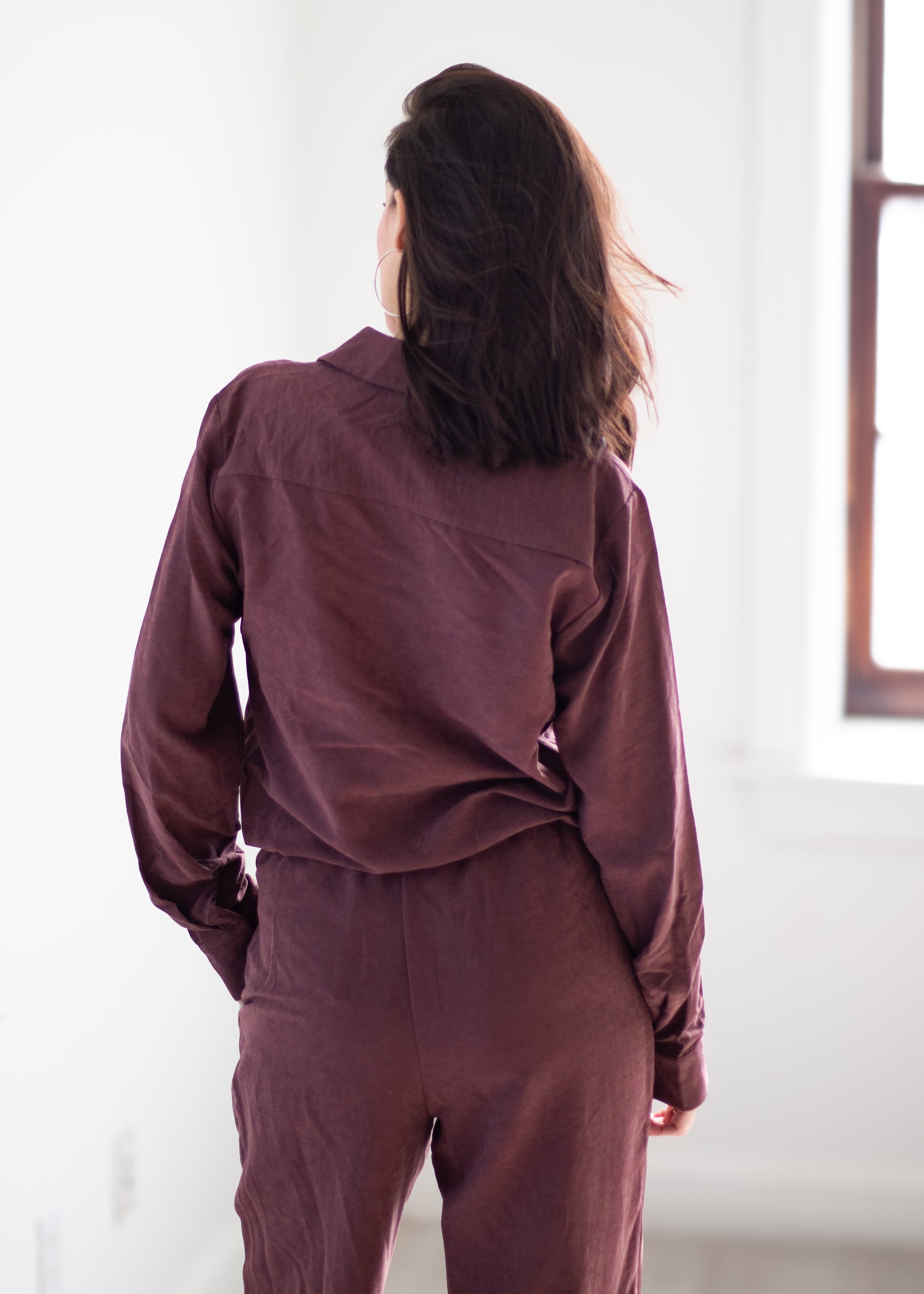 A woman viewed from behind wearing a matching burgundy long-sleeve button-up shirt and trousers set in a bright, minimalist room.