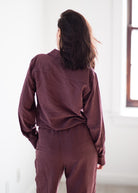 A woman viewed from behind wearing a matching burgundy long-sleeve button-up shirt and trousers set in a bright, minimalist room.