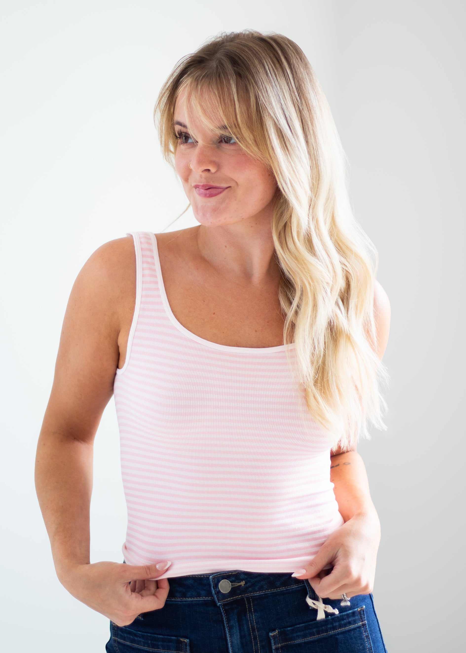 A woman with blonde hair wearing a light pink and white striped ribbed tank top and dark blue jeans, posing against a light gray studio background.