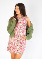 A woman modeling a sleeveless floral mini dress with a green knit cardigan draped off her shoulders, set against a plain white studio background.