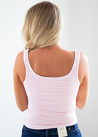 A woman viewed from the back wearing a light pink and white horizontally striped tank top and blue jeans against a plain light gray background.