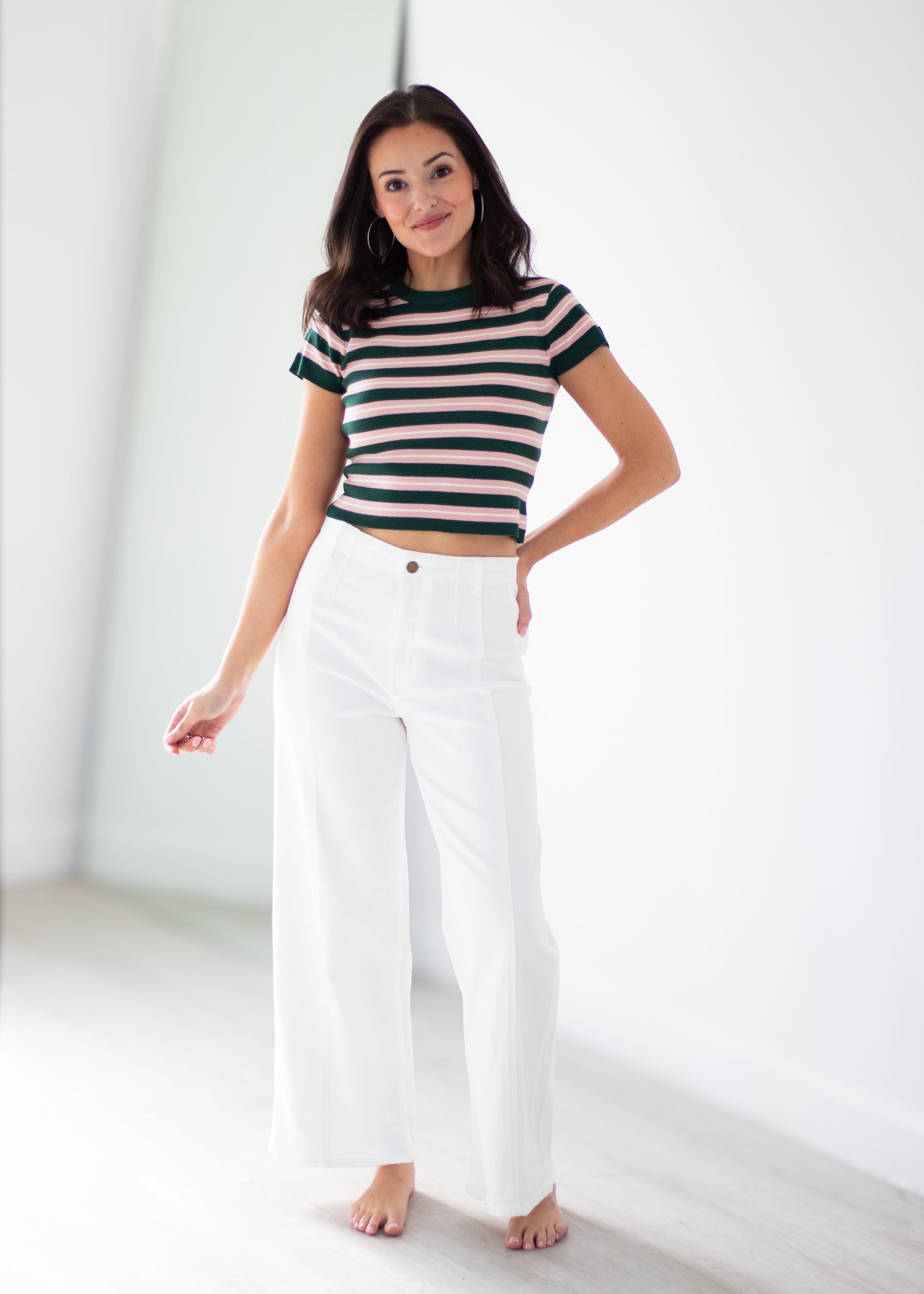 A woman modeling a short-sleeve striped crop top with dark green and beige horizontal stripes, paired with high-waisted white wide-leg pants, standing in a bright, minimalist white room.