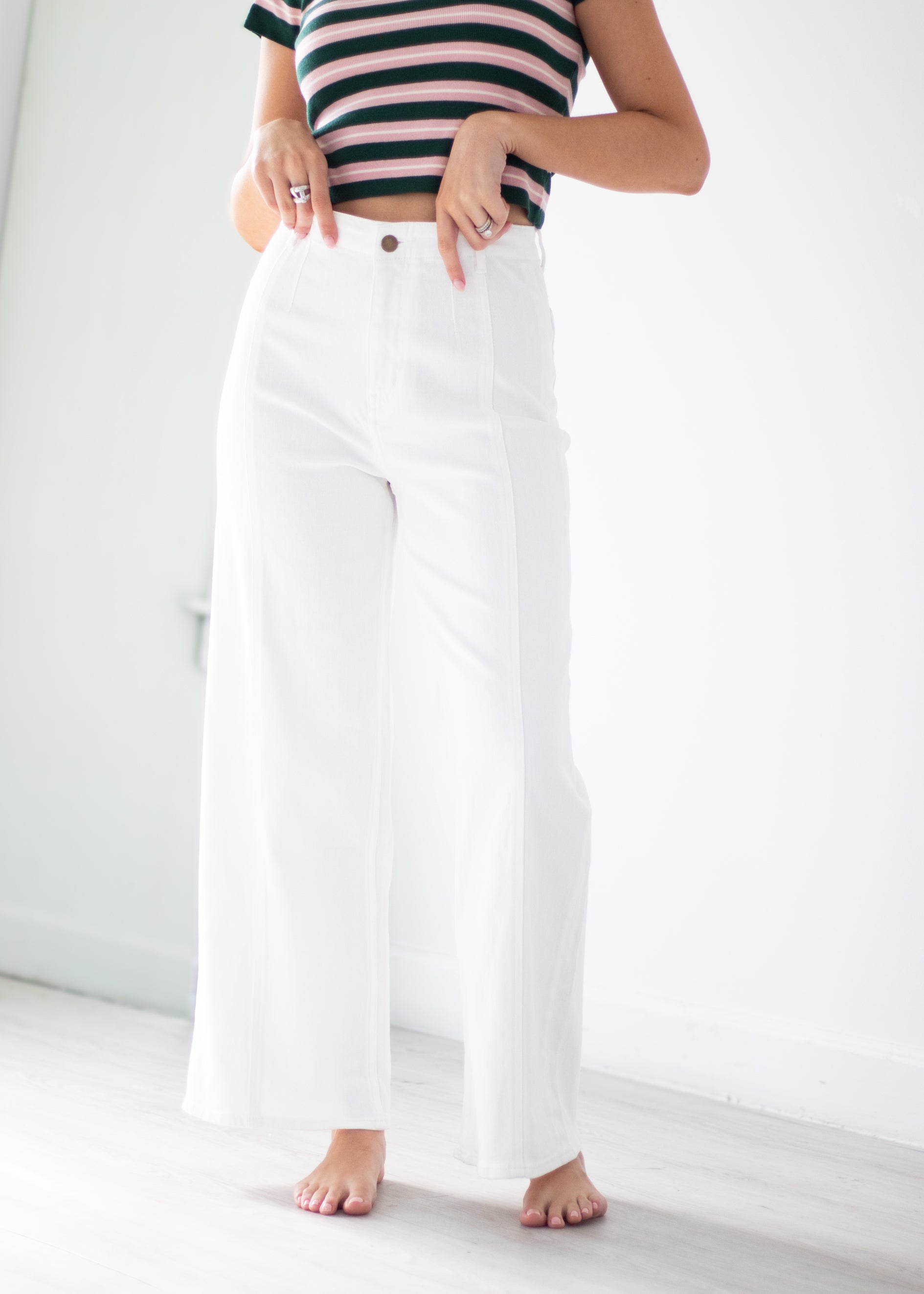 A person wearing high-waisted, wide-leg white pants and a navy and white striped crop top, standing barefoot in a bright, minimalist room with white walls and light wood flooring.