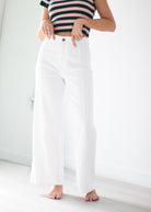 A person wearing high-waisted, wide-leg white pants and a navy and white striped crop top, standing barefoot in a bright, minimalist room with white walls and light wood flooring.