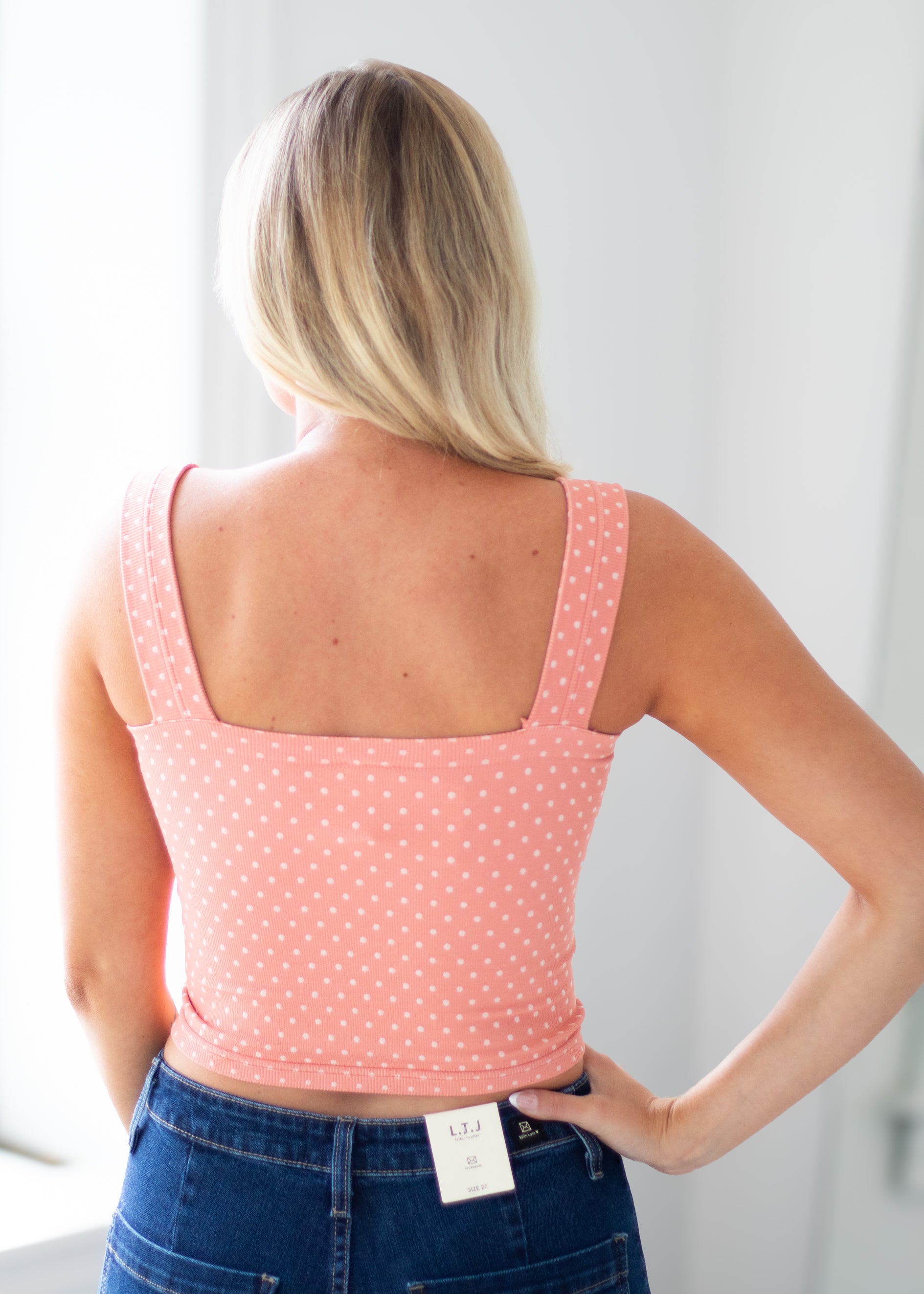 A woman viewed from behind wearing a coral pink tank top with small white polka dots and blue jeans, standing in a bright indoor setting.