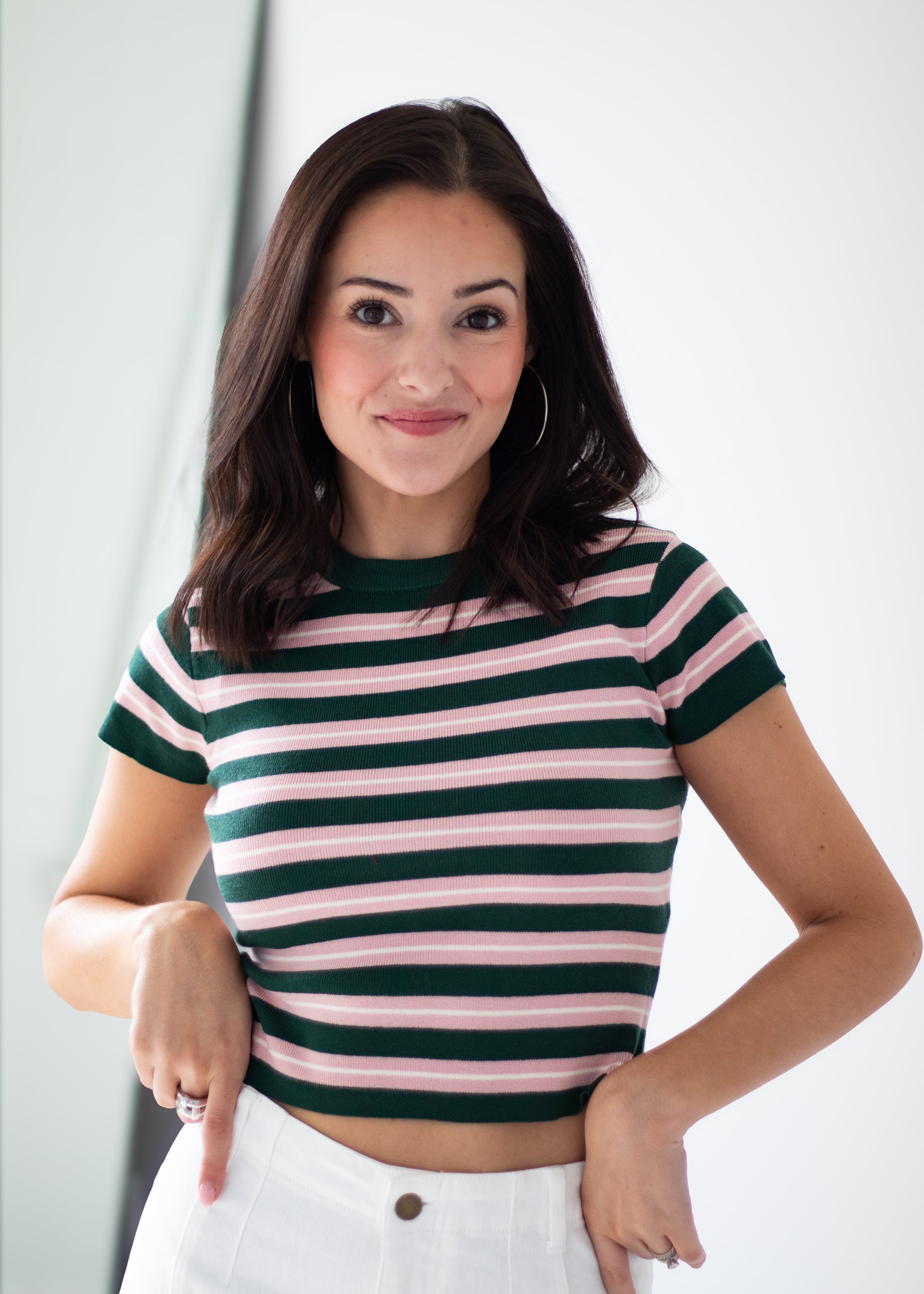A woman wearing a short-sleeve crop top with horizontal dark green and light pink stripes, paired with white pants, standing against a light gray background.
