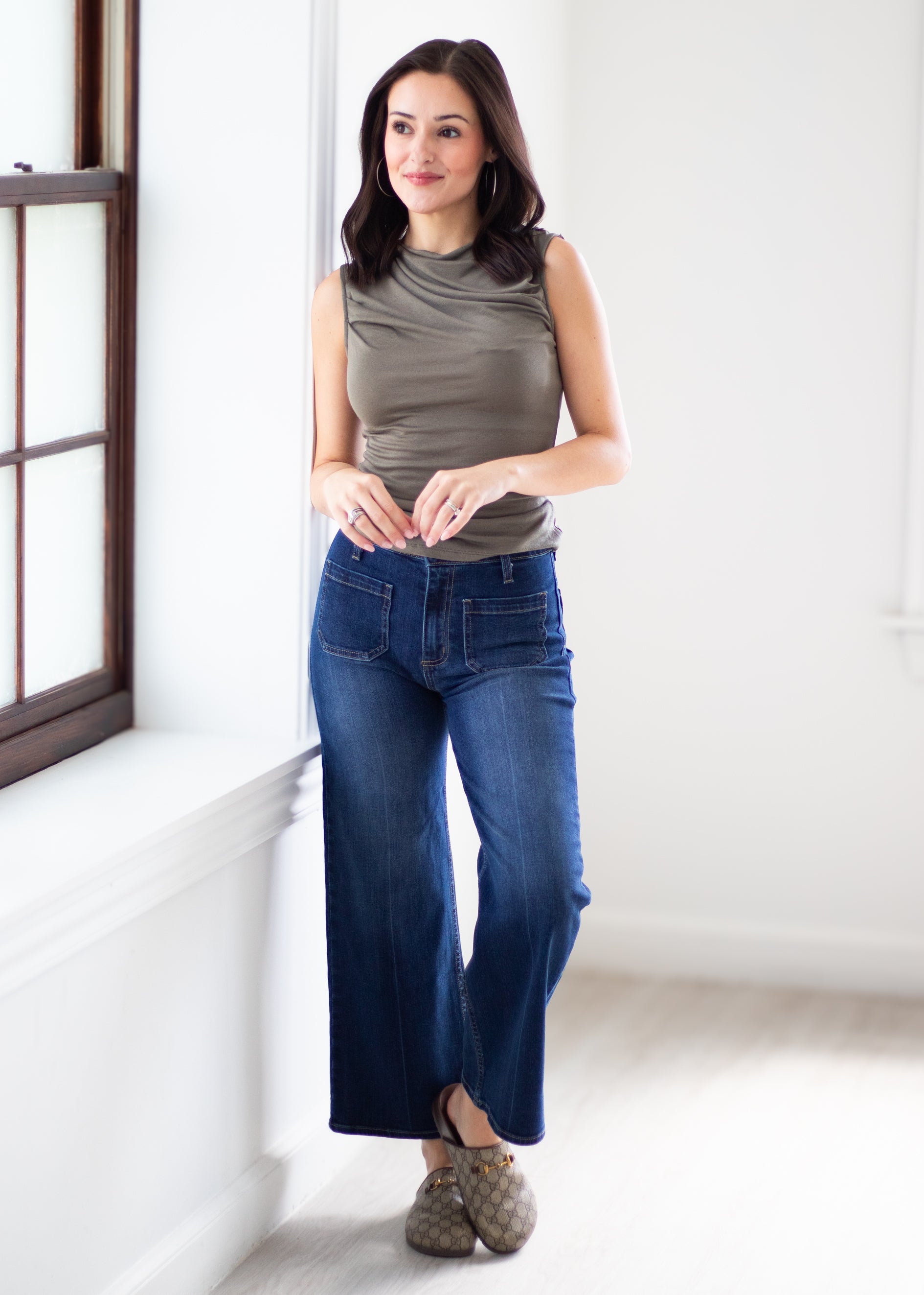 A woman stands in a bright white room wearing a sleeveless taupe top and dark blue wide-leg cropped jeans with brown shoes.