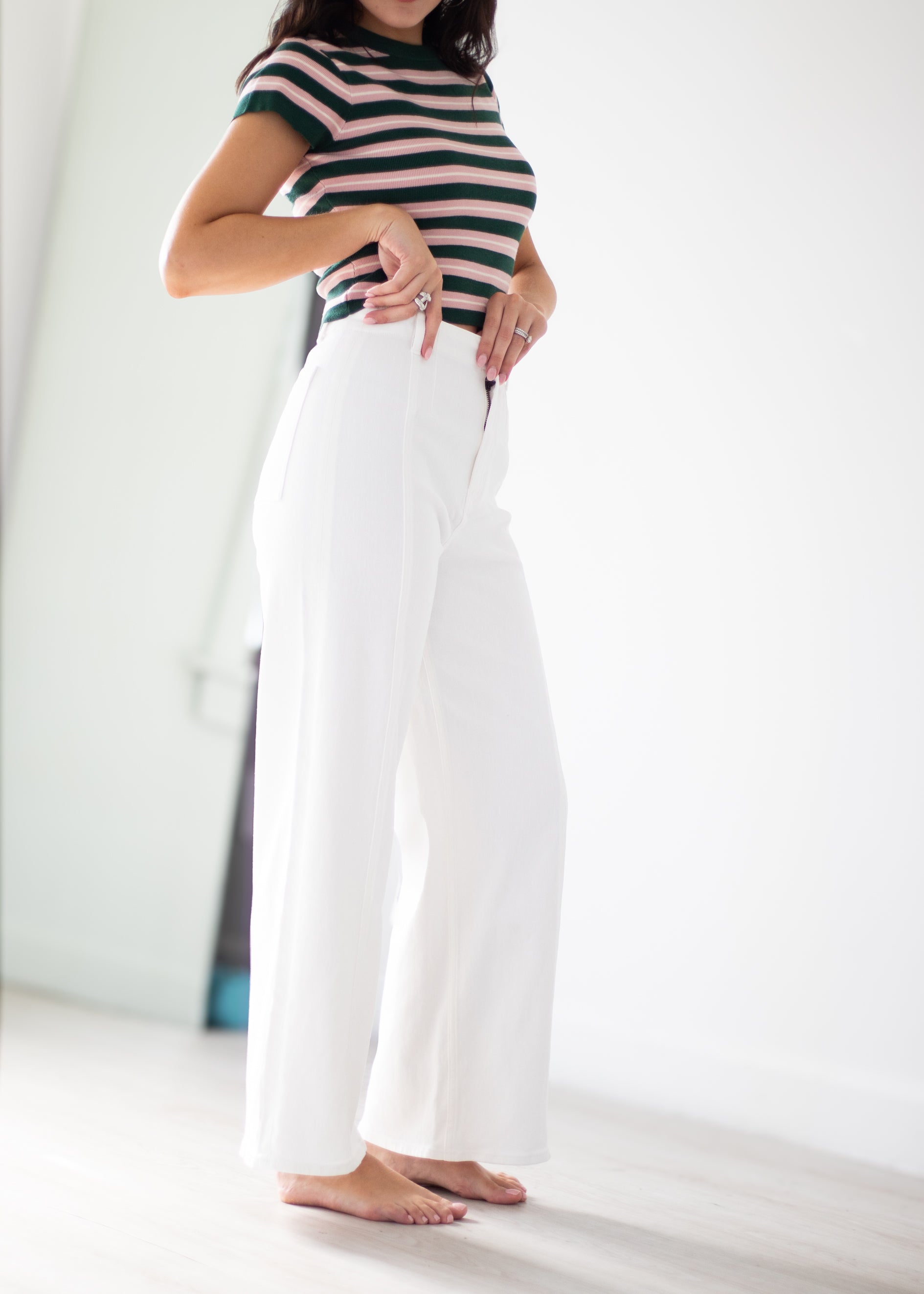 A woman wearing a black and white horizontally striped short-sleeve top and high-waisted, wide-leg white trousers, standing barefoot in a bright, minimalist room.