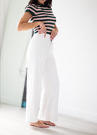 A woman wearing a black and white horizontally striped short-sleeve top and high-waisted, wide-leg white trousers, standing barefoot in a bright, minimalist room.