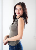A woman with long brown hair is wearing a sleeveless taupe-gray draped tank top and blue jeans, posing in a brightly lit indoor setting.