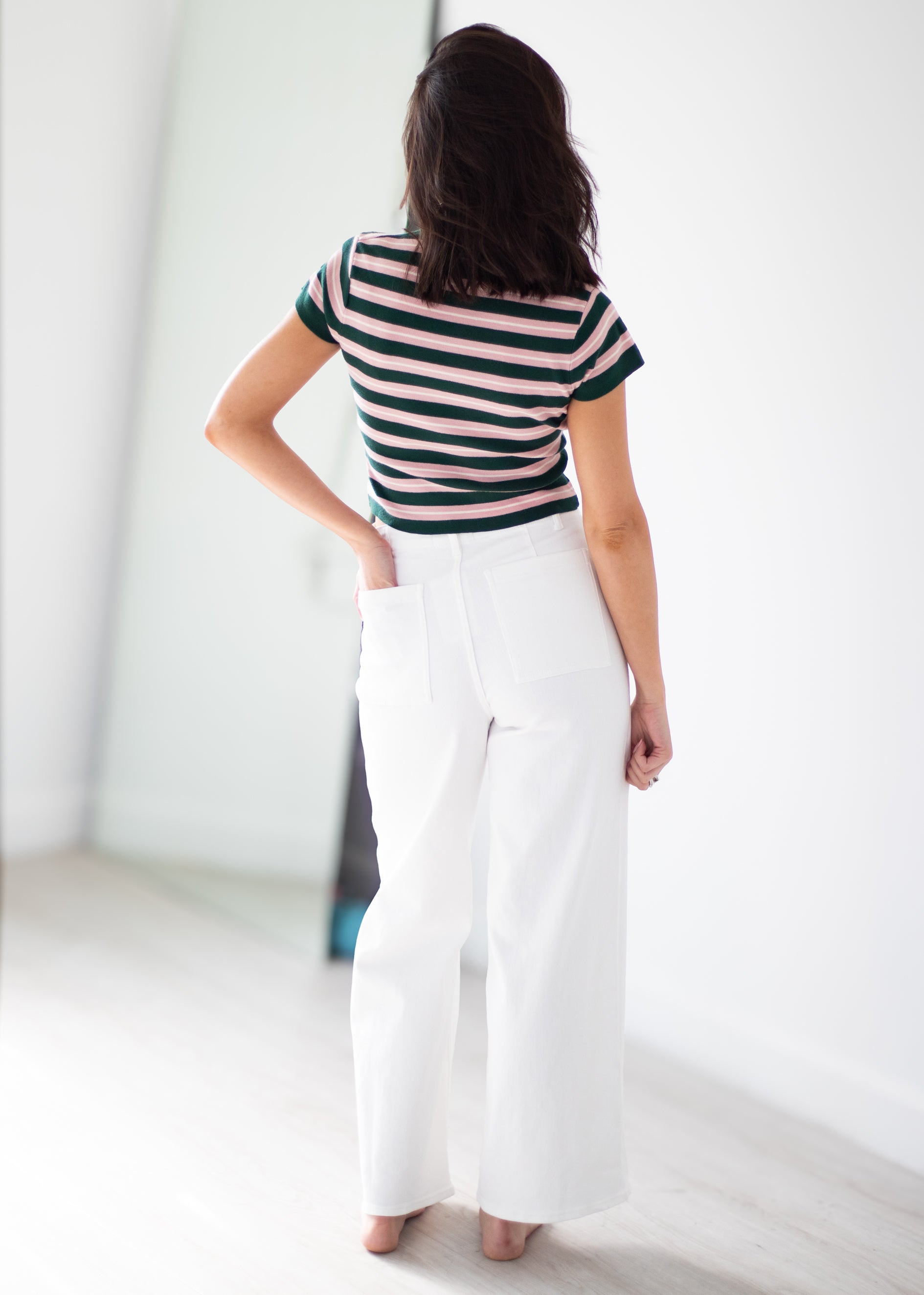A woman shown from behind wearing a short-sleeve striped top with dark green and light pink horizontal stripes, paired with high-waisted white wide-leg pants, standing in a bright minimalist room.