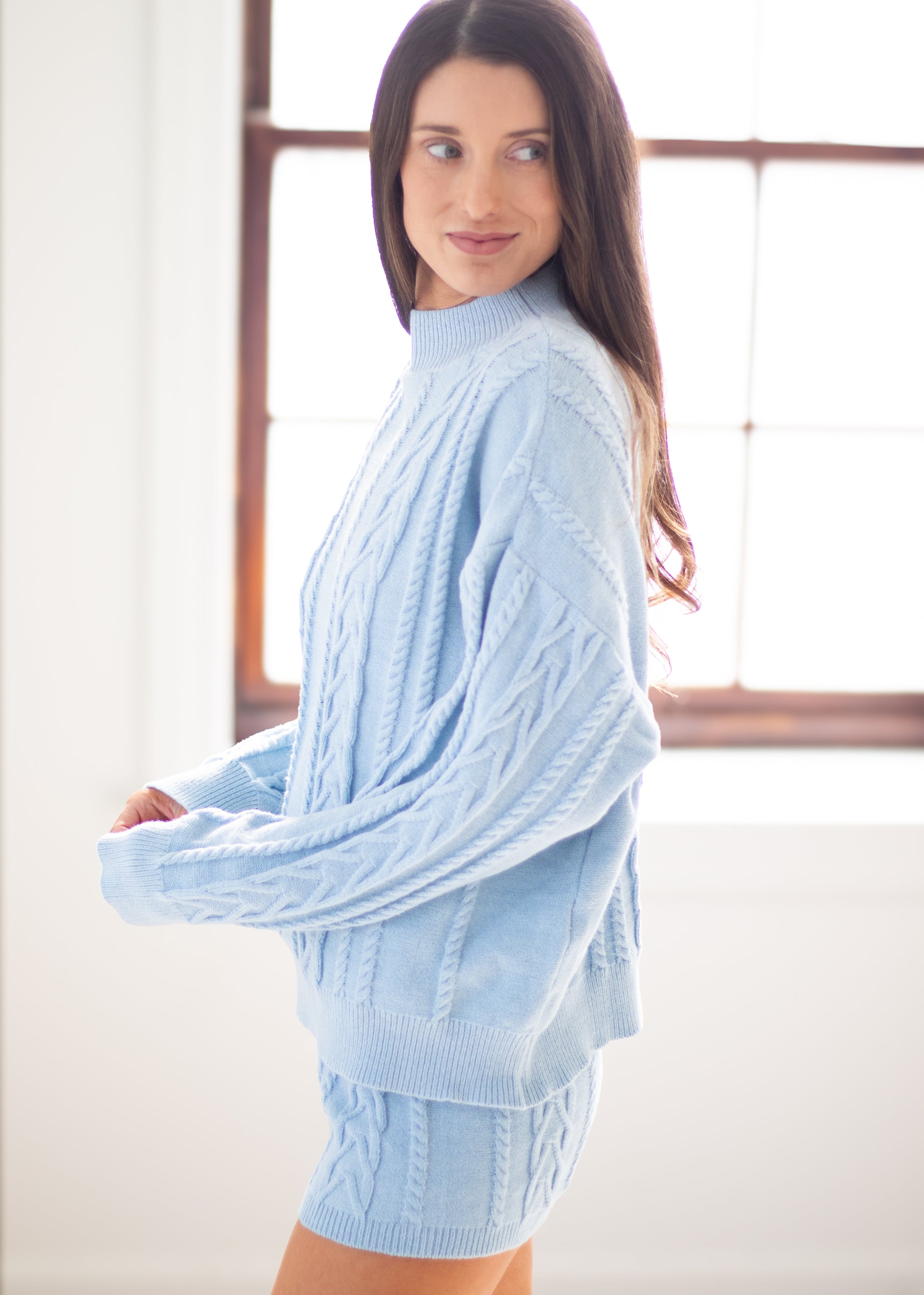 A woman wearing a light blue cable-knit mock-neck sweater and matching knit shorts, standing in a bright room with a window in the background.