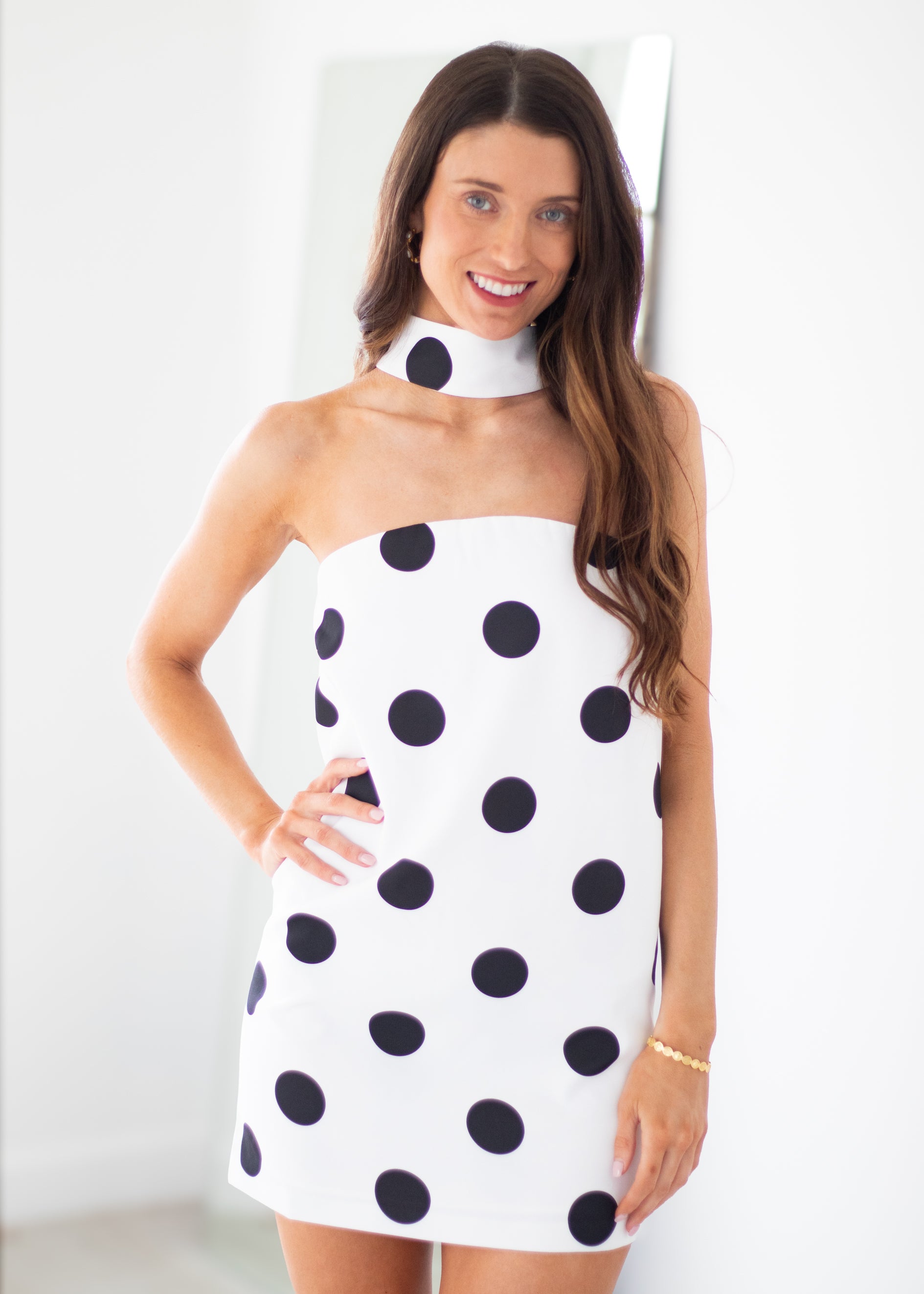 A woman modeling a white halter-style mini dress with large black polka dots, standing against a bright white indoor background.