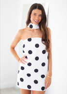 A woman modeling a white halter-style mini dress with large black polka dots, standing against a bright white indoor background.