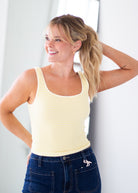 A woman wearing a pale yellow and white horizontally striped ribbed tank top and dark blue jeans, posing in a brightly lit indoor setting.