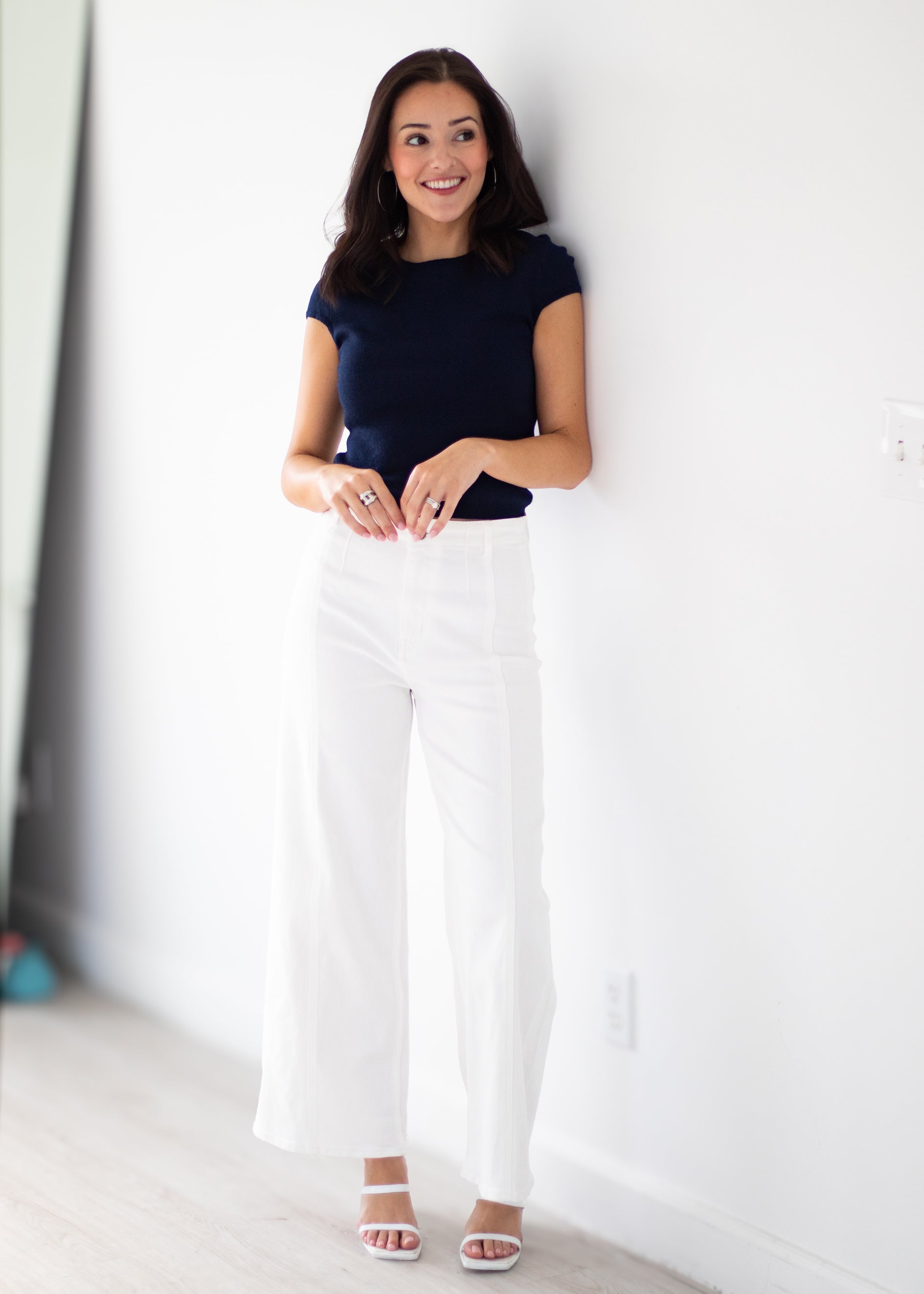 A woman stands against a white wall wearing a navy blue short-sleeve top, white wide-leg cropped pants, and white heeled sandals.