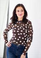 A woman wearing a brown long-sleeve mock neck top with large white polka dots, paired with blue jeans, standing against a white studio background.